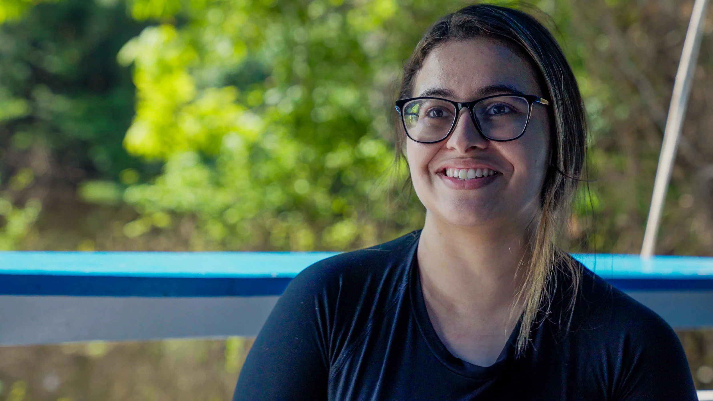 Smiling person wearing glasses, in front of a blurred green foliage background.