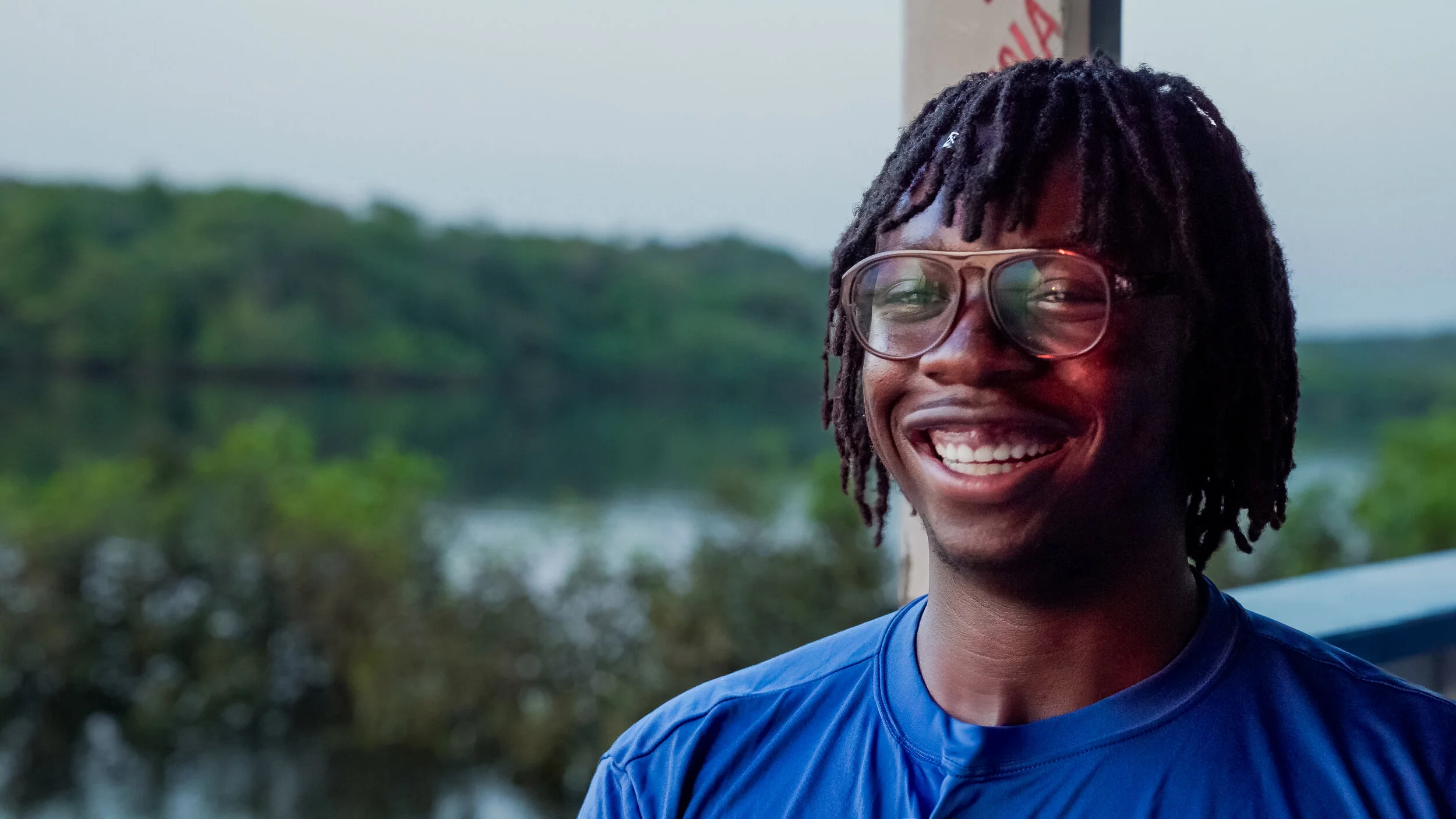 Person with glasses smiling outdoors near water and trees.