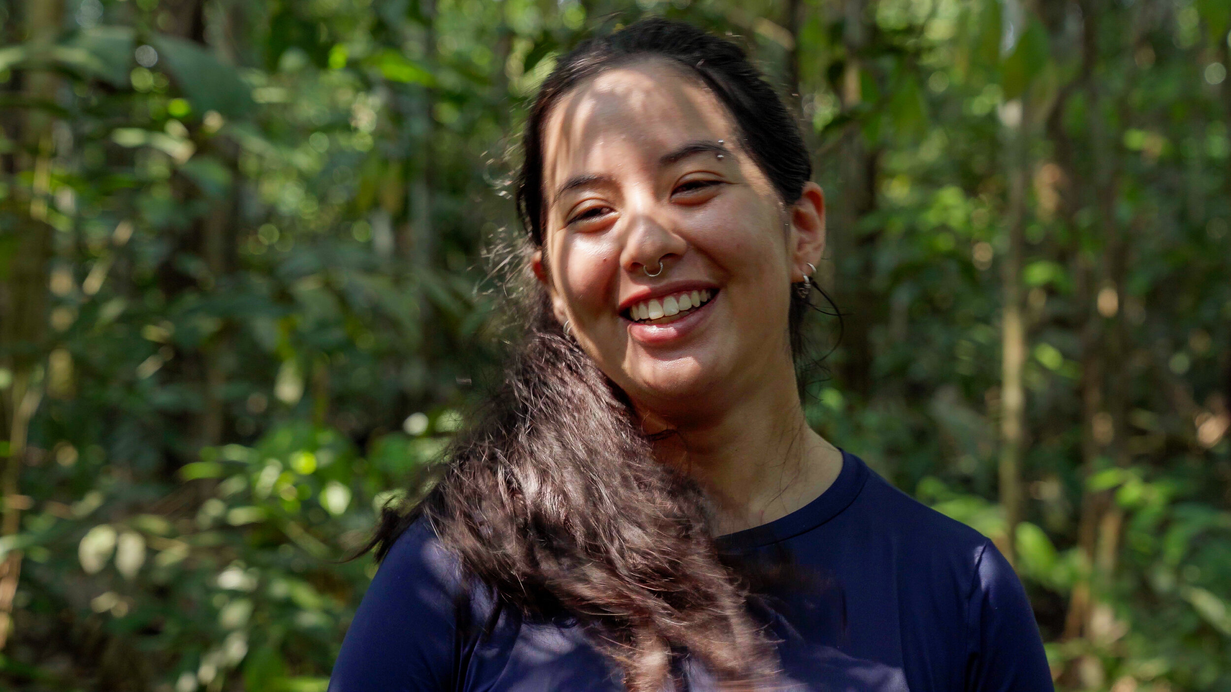 A woman smiling joyfully in a sunlit forest, wearing a dark blue shirt with long dark hair and a nose ring.