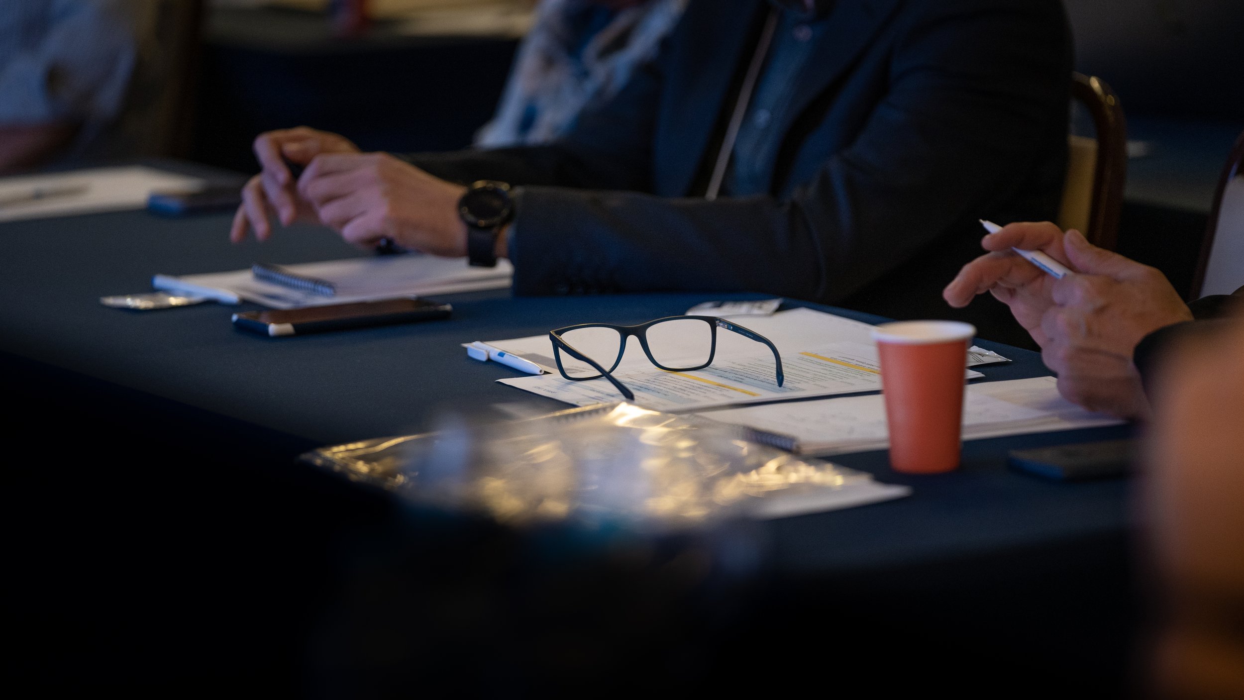 Close-up of a table at a meeting with eyeglasses, papers, a pen, smartphone, and a red disposable cup.