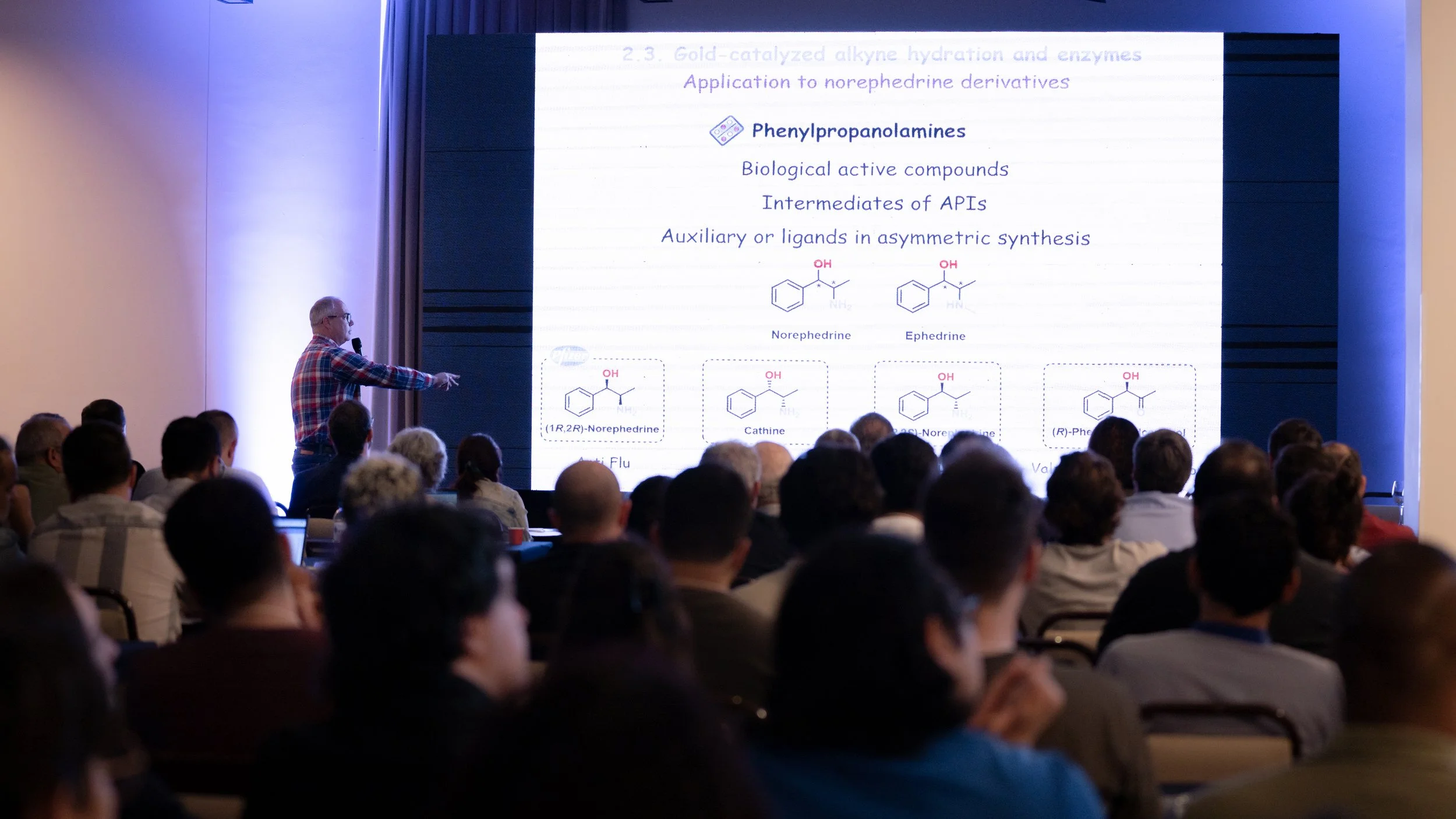 A speaker presenting a chemical lecture at a conference with a PowerPoint slide on phenylpropanolamines to an audience.