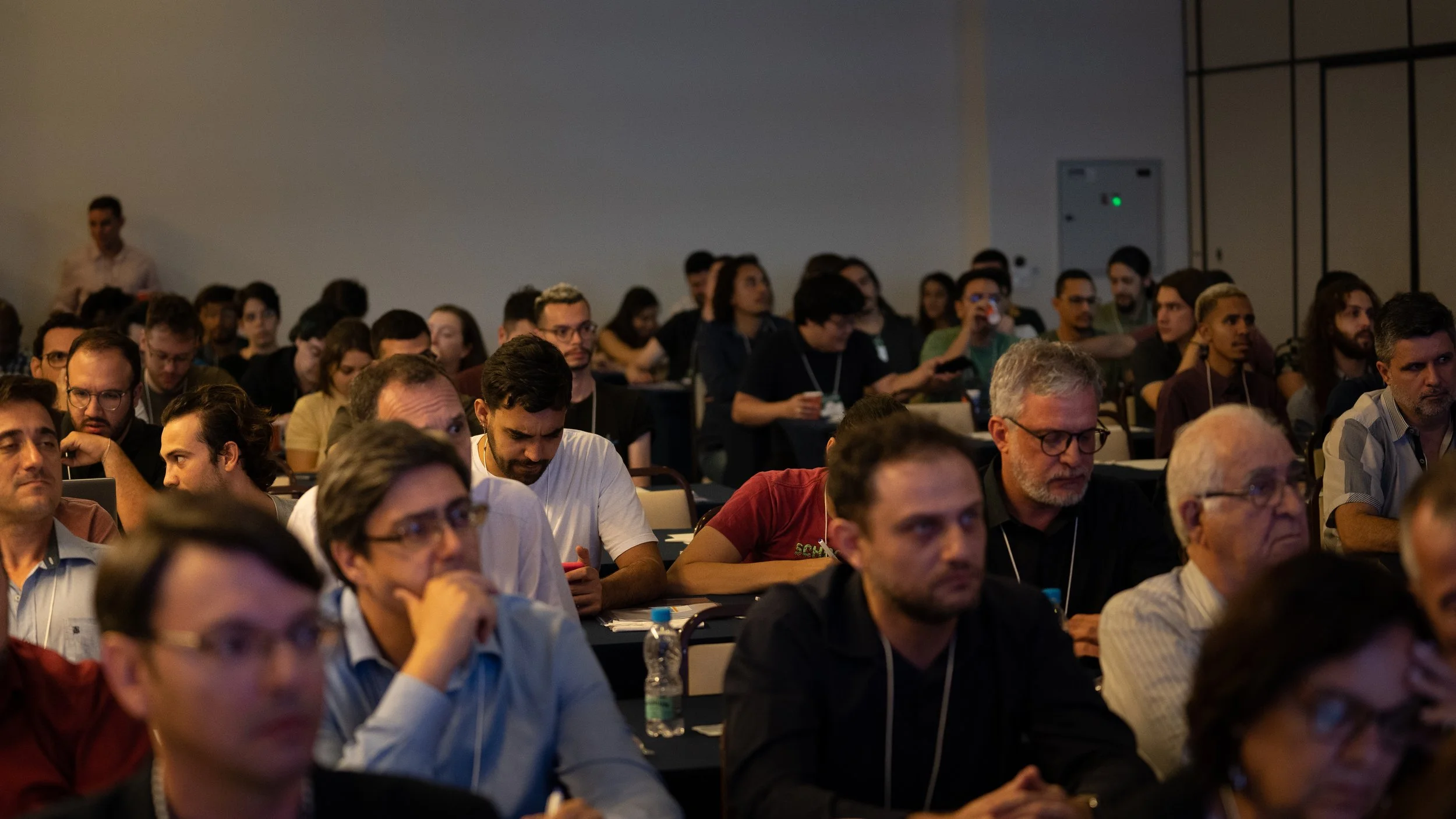 Audience attending a conference in a dimly lit room, with a focus on people listening and taking notes.