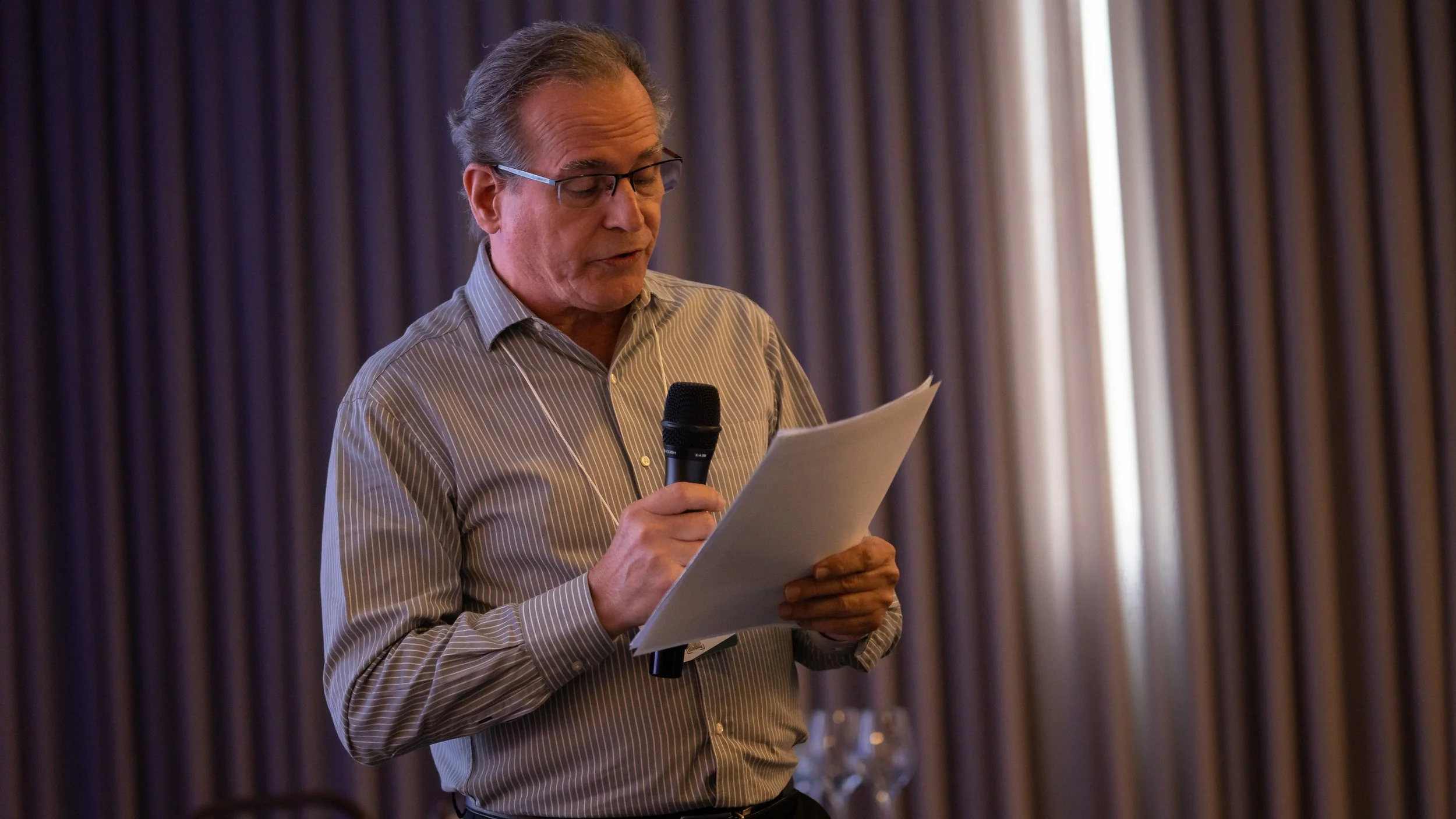 Man in striped shirt holding a microphone and reading from a sheet of paper.