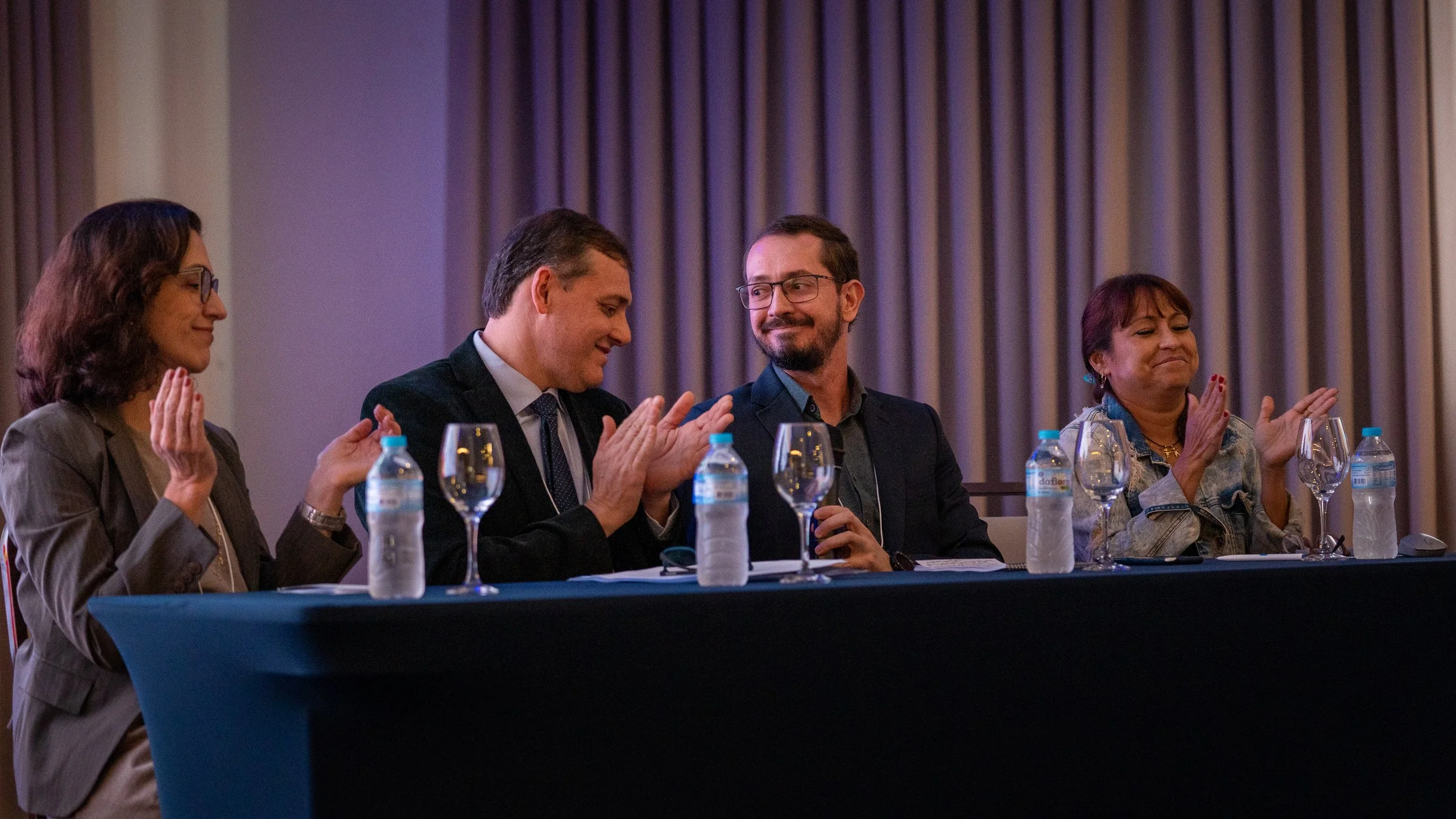 A panel of four people sitting at a table clapping, with water bottles and empty wine glasses in front of them.