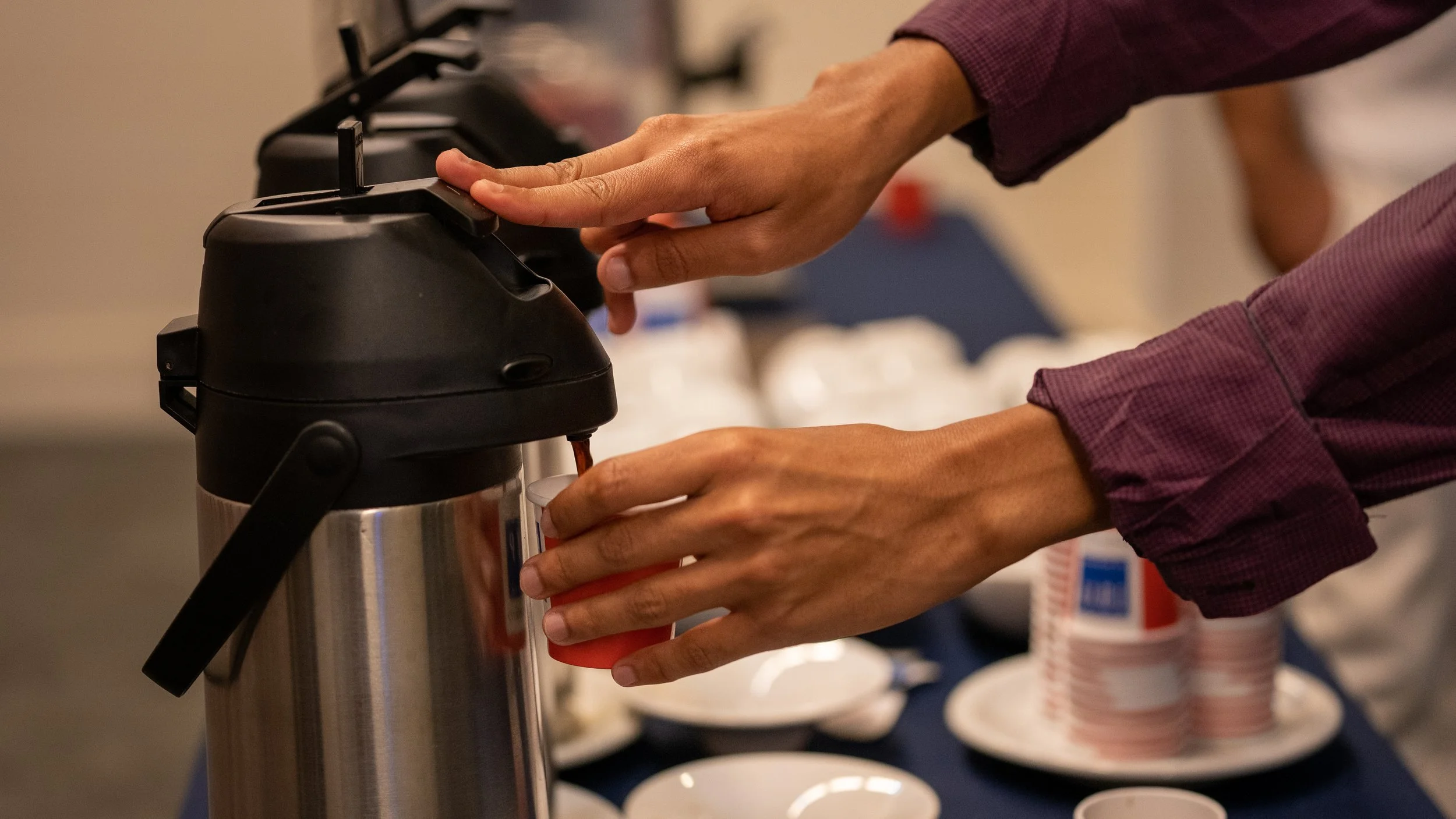 Person pouring coffee into a disposable cup from an airpot dispenser, with stacks of cups and saucers in the background.
