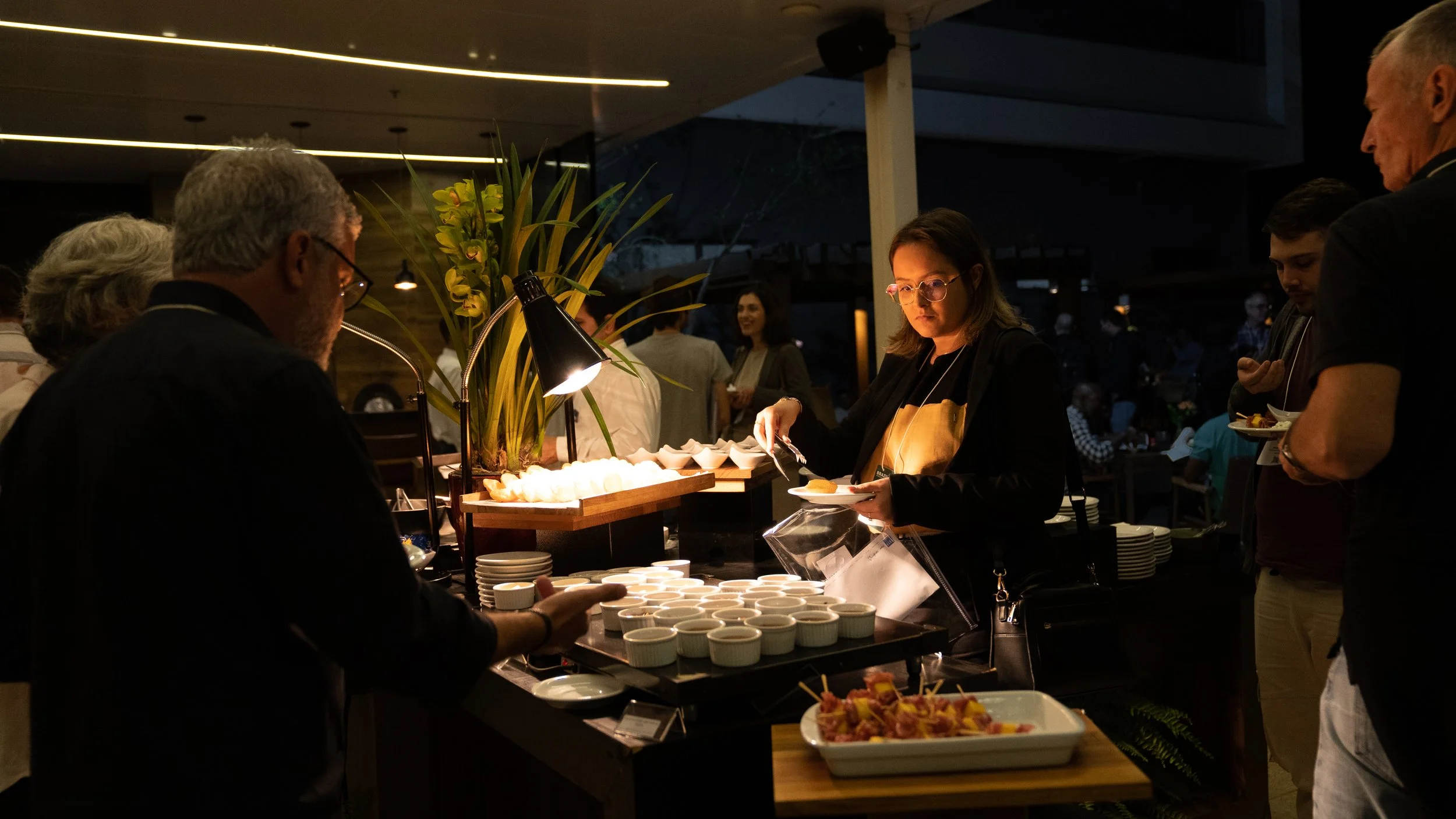 People at a buffet table selecting food in a dimly lit indoor setting.
