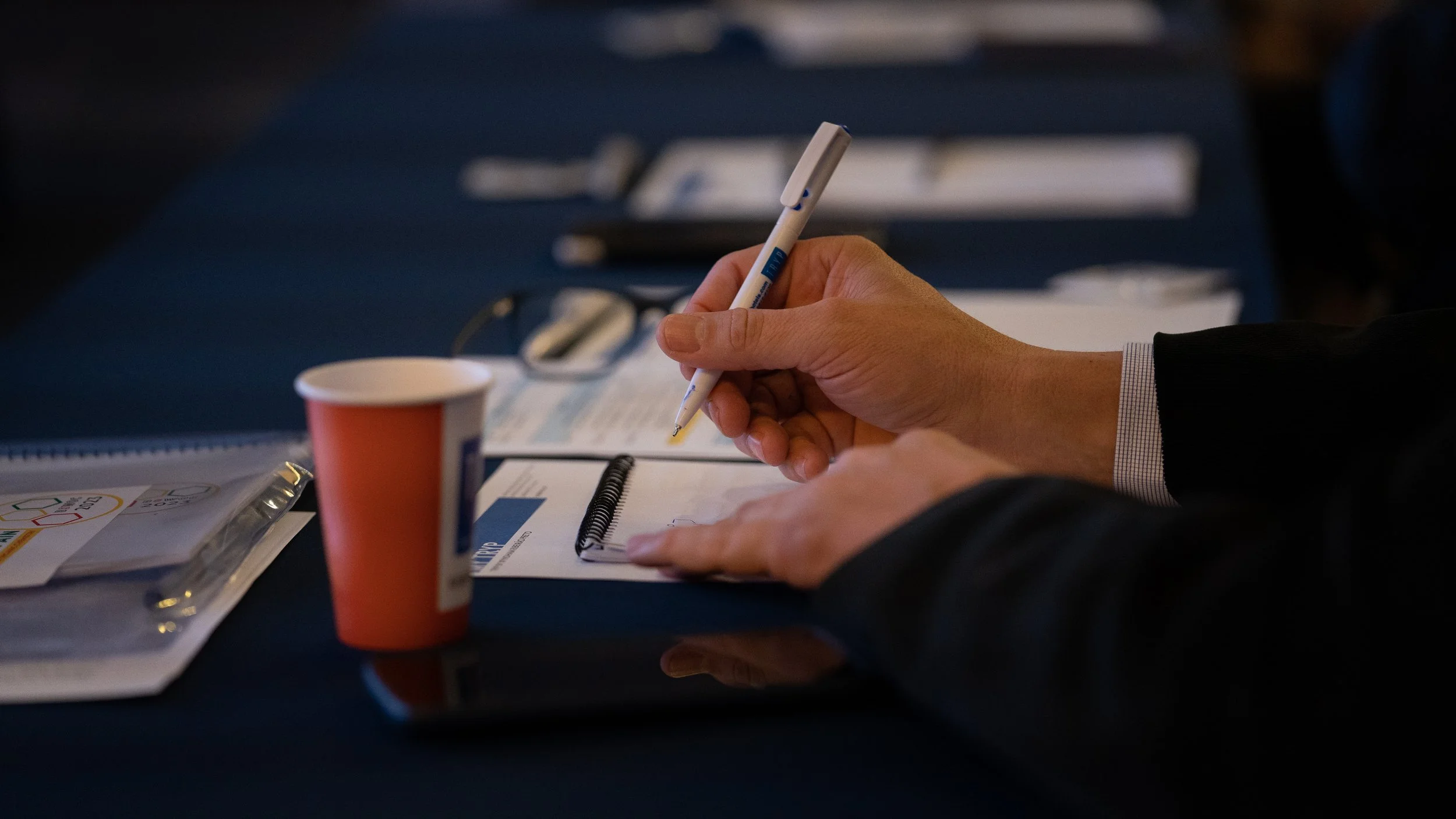 Person's hand writing on a notepad with a pen at a conference table, accompanied by a red paper cup and eyeglasses.