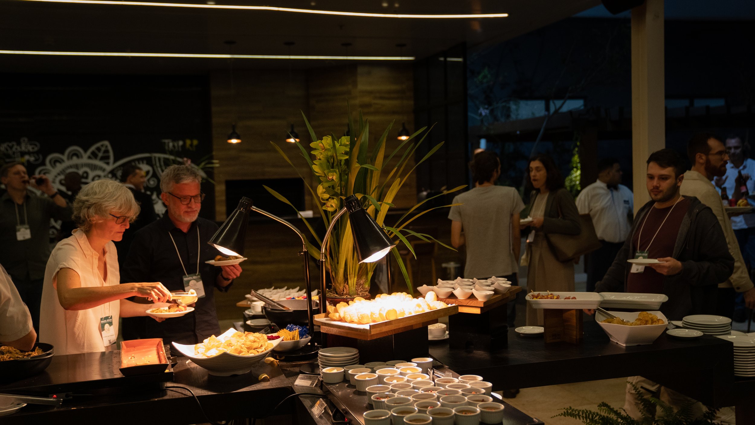 People serving themselves food at a buffet during an indoor event, with various dishes and decorative lighting.