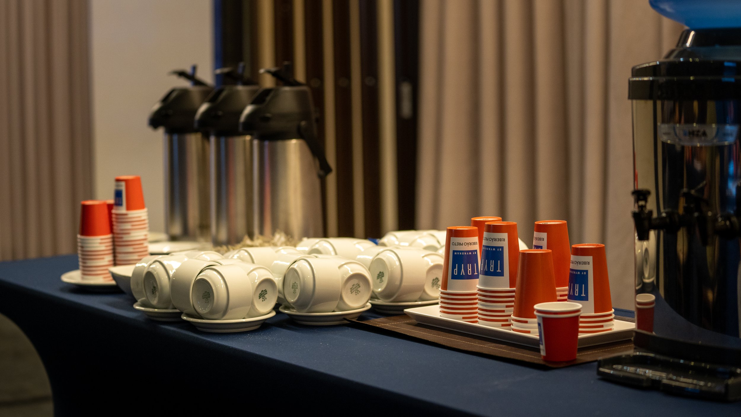 Coffee station with stacked paper cups, ceramic cups on saucers, thermal carafes, and a water dispenser on a table.