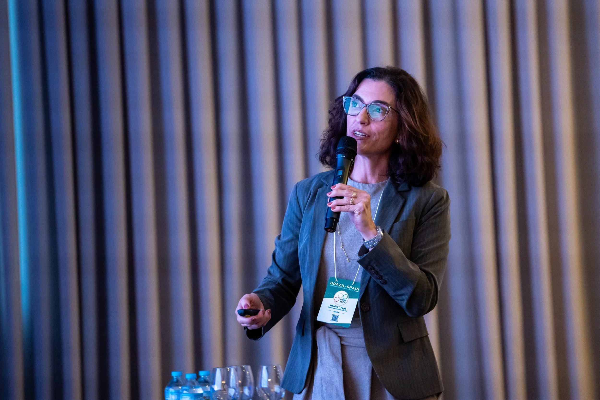 Woman speaking into microphone at an event, with a badge and holding a remote, standing by a table with water bottles.