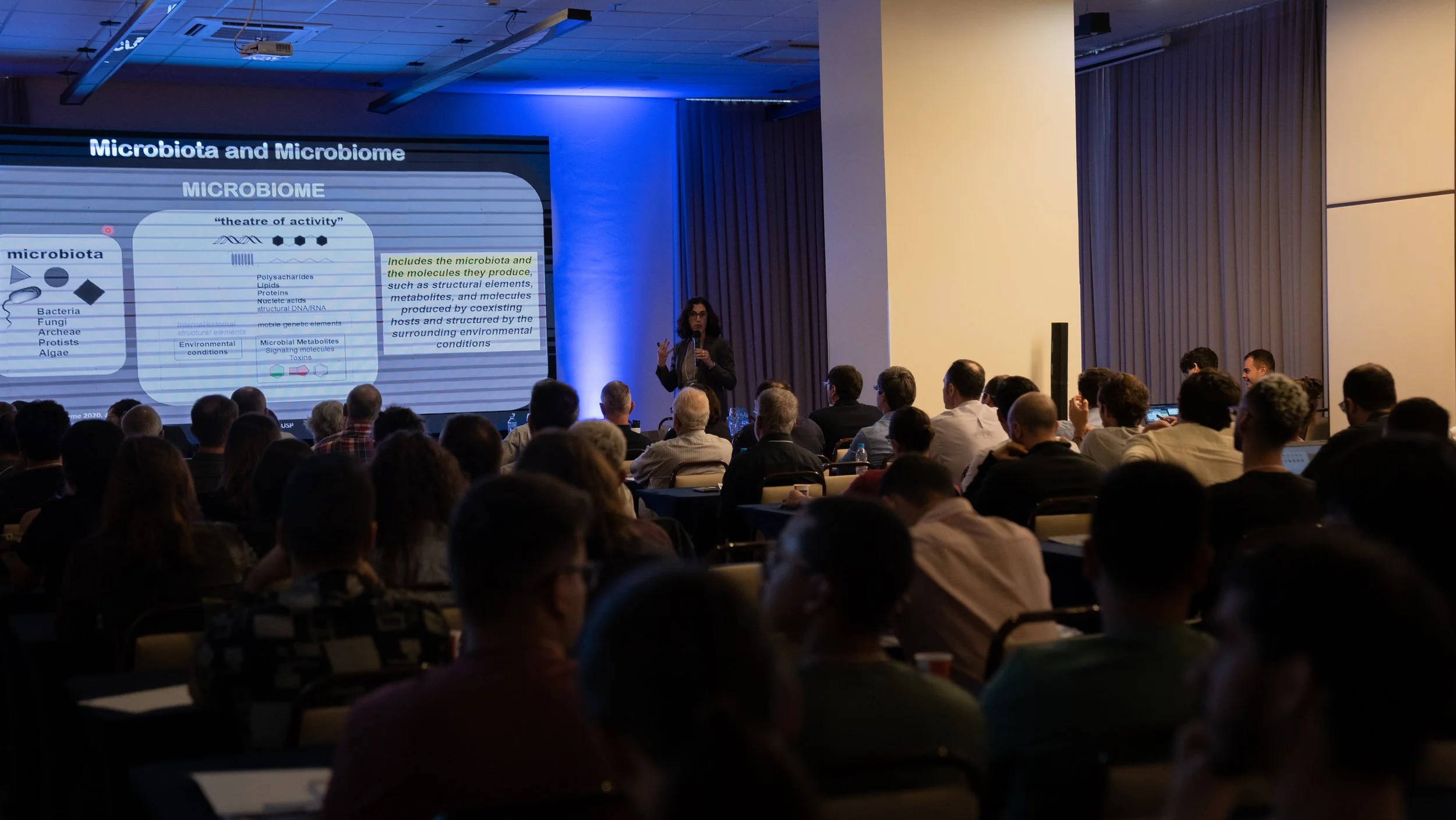 Audience at a conference listening to a speaker with a presentation on microbiota and microbiome.