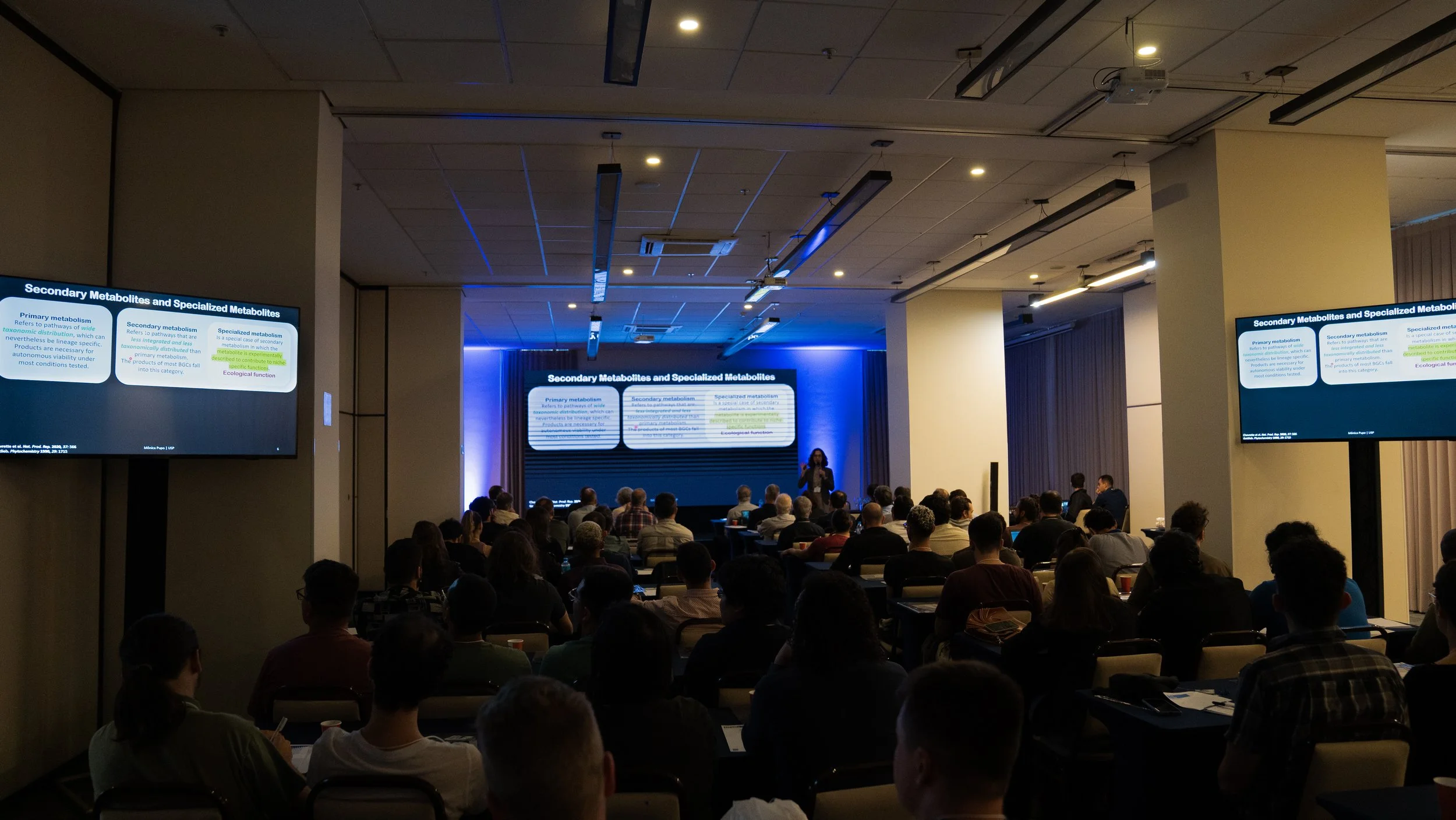 Audience attending a presentation on secondary metabolites and specialized metabolites, with slides displayed on screens around the room.