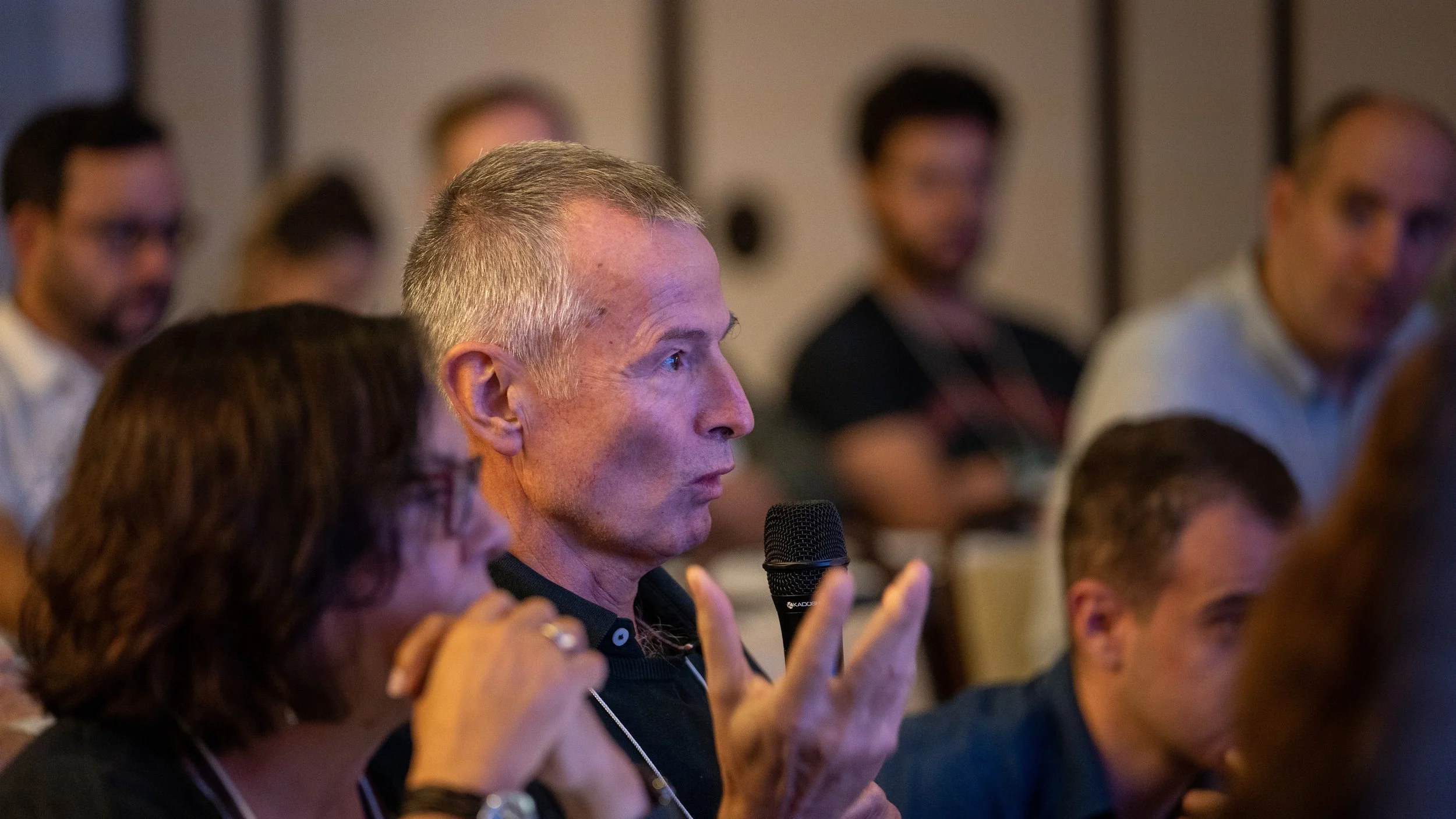 Man speaking into a microphone during a conference, surrounded by an audience seated in a room.