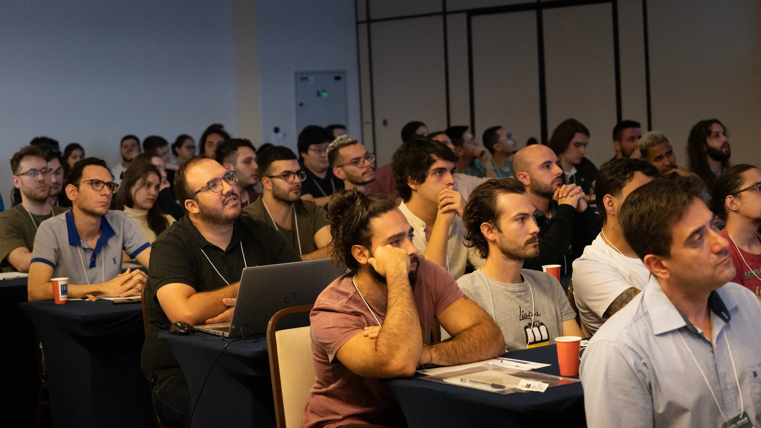 Audience attentively listening to a presentation at a conference, with people sitting at tables, some with laptops and notebooks, in a dimly lit room.