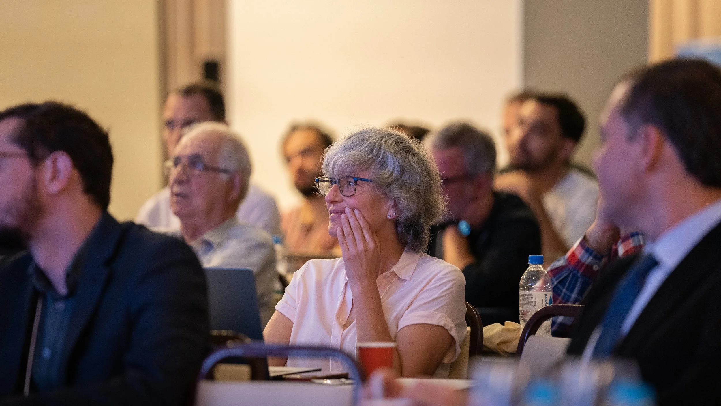 Audience listening intently during a conference or presentation, featuring a woman with gray hair and glasses in the foreground, seated among others in a brightly lit room.
