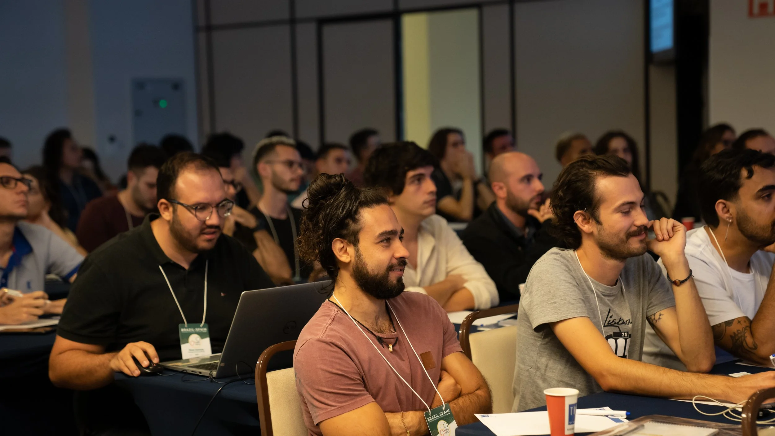A diverse group of people attending a conference, sitting at tables, looking engaged. Some are using laptops, while others are listening intently. They appear to be in a classroom or seminar setting.