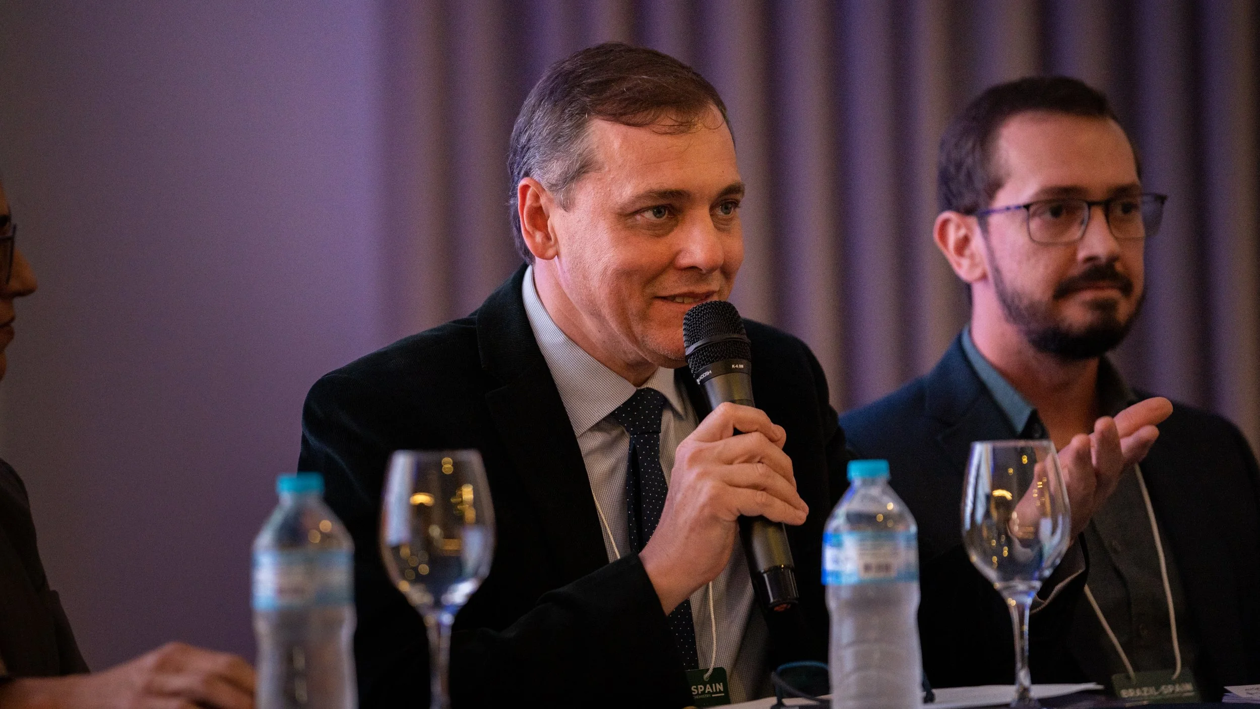Two men sitting at a table, one speaking into a microphone, with water bottles and wine glasses in front of them.