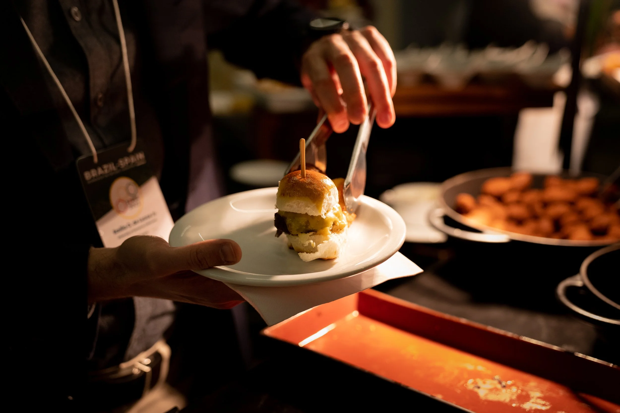 Person holding a plate with a slider sandwich being served with tongs at an event.