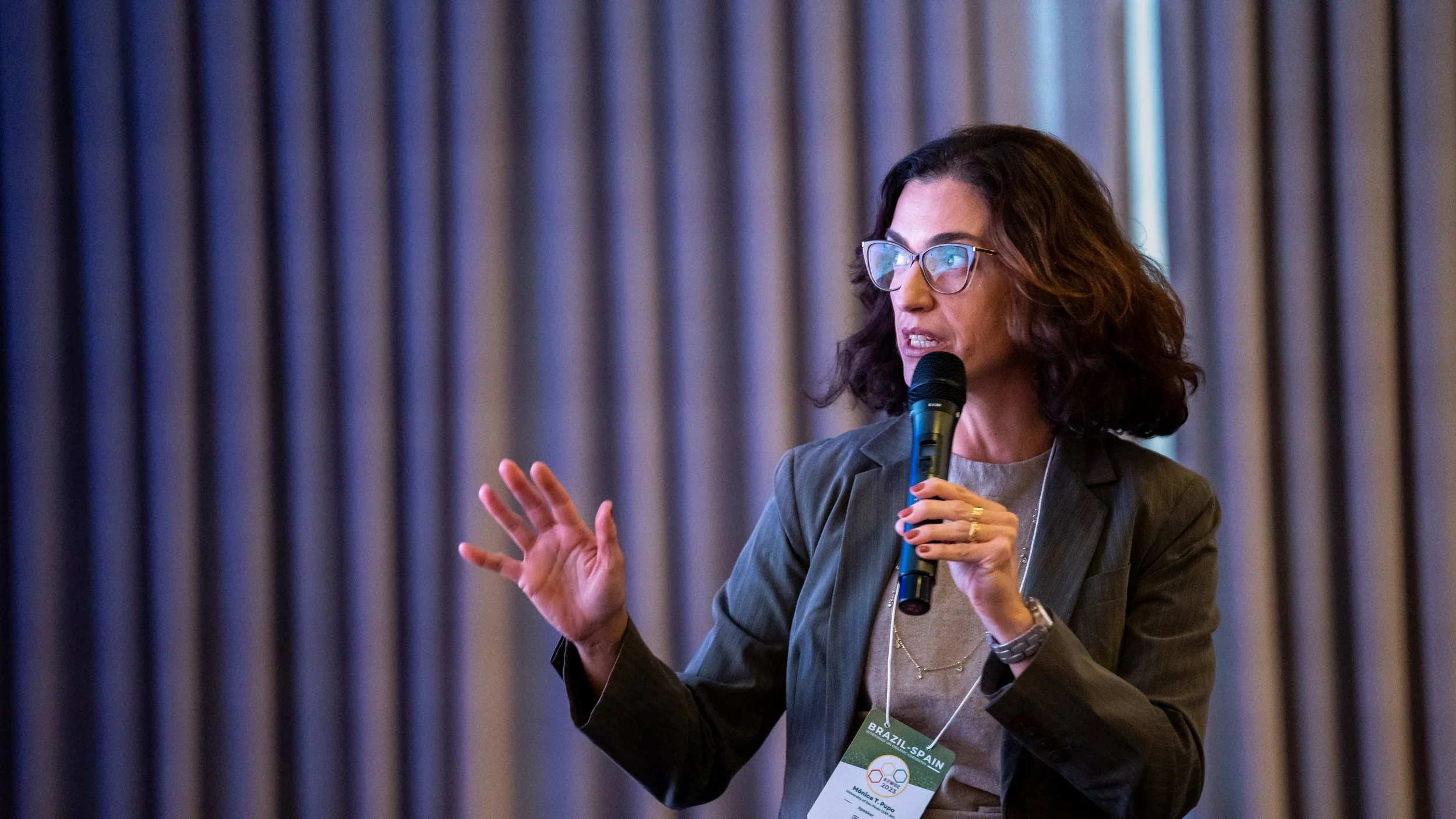Person speaking into a microphone at a conference, wearing glasses and a name tag.