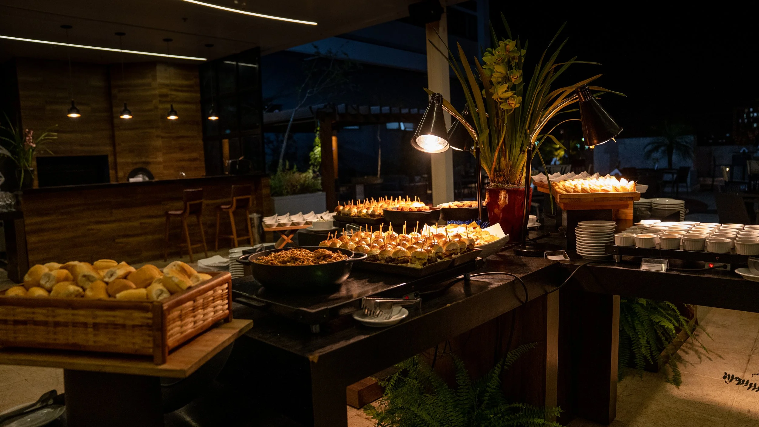 Buffet setup with assorted foods, including mini sandwiches on skewers, a bowl of noodles, a basket of rolls, stacks of plates, cups on a warmer, and decorative flowers. Background features a wooden bar area with pendant lighting.
