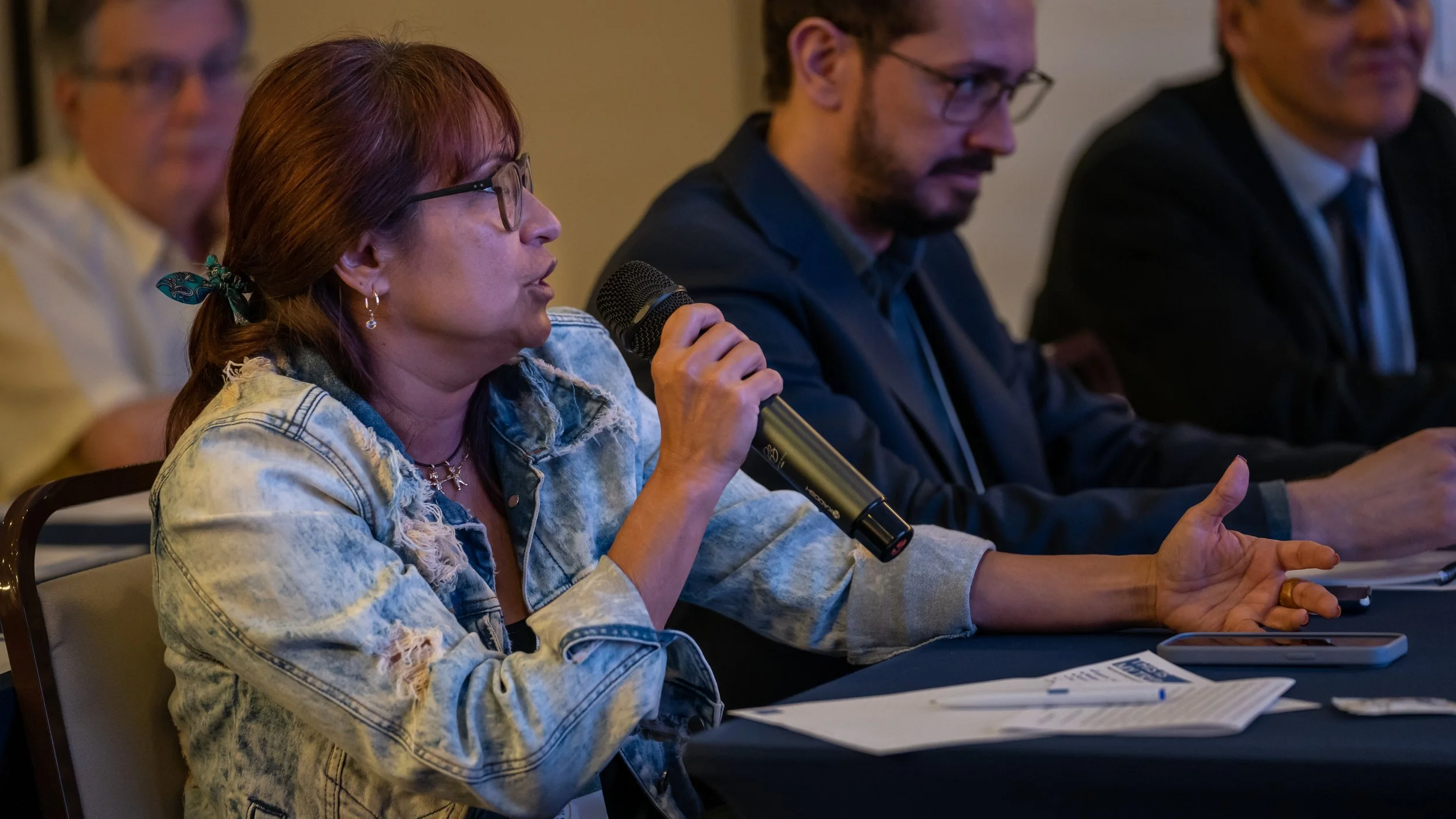 Woman speaking into a microphone during a meeting, wearing glasses and denim jacket, with two men in suits seated nearby.