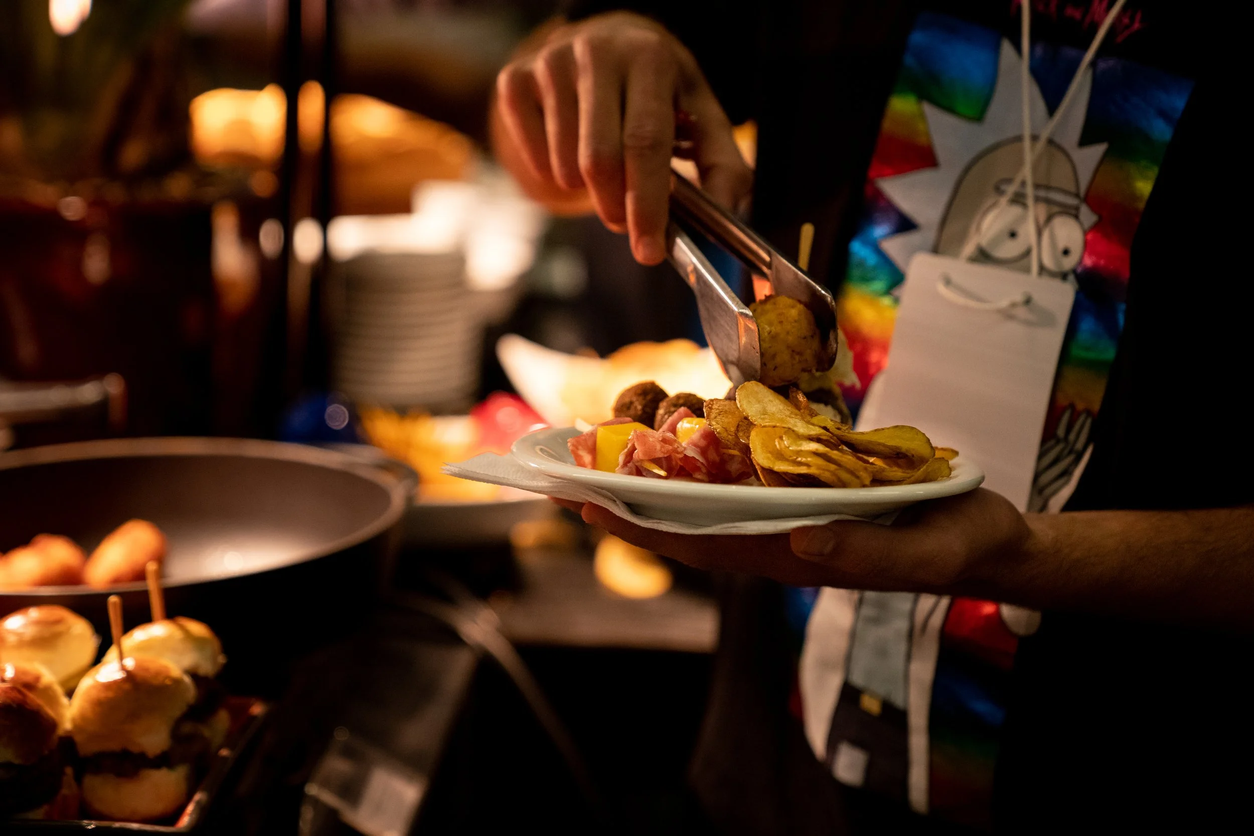Person serving appetizers onto a plate with tongs, including potato chips and sliders, wearing a colorful cartoon-themed shirt.