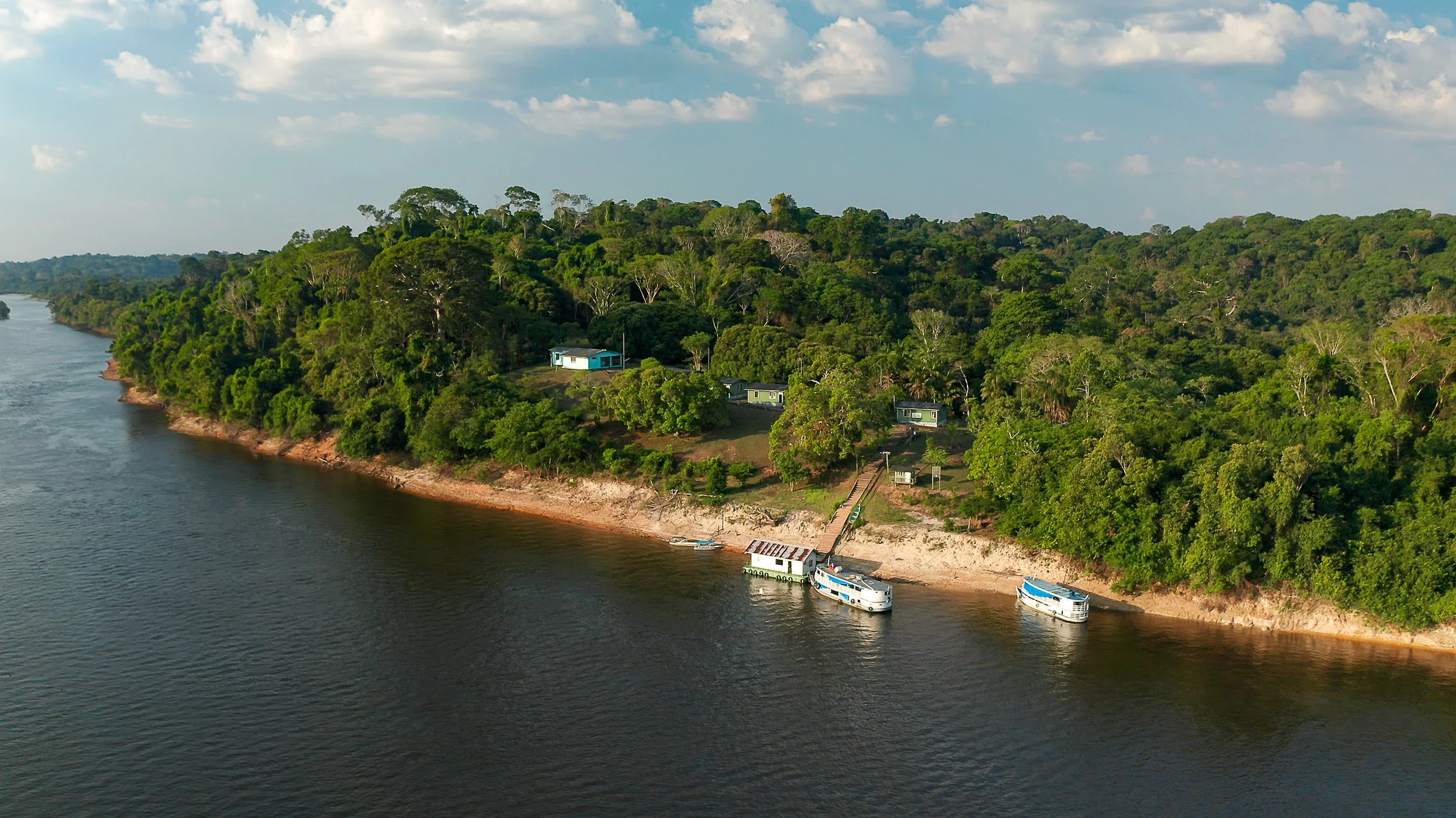 Aerial view of a river with boats docked along a forested shoreline, surrounded by dense greenery and some buildings visible among the trees, under a partly cloudy sky. TheICMBio Trombetas - Base avançada Tabuleiro Research Station in the Amazon