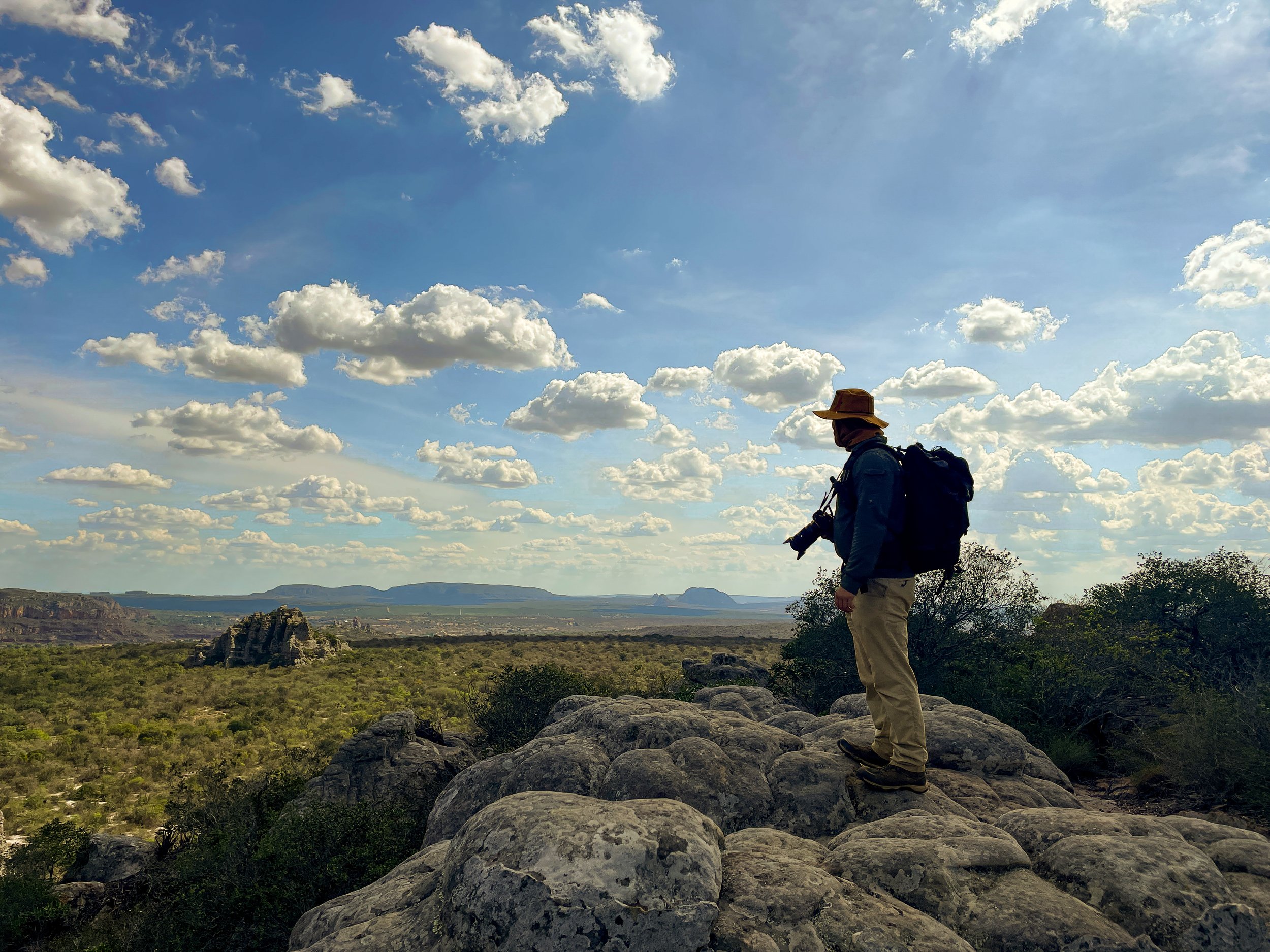 A person, Basil Minder wearing a hat and backpack stands on a rocky outcrop, holding a camera. The background features a vast landscape of caatinga with scattered clouds in a blue sky, and distant mountains on the horizon.