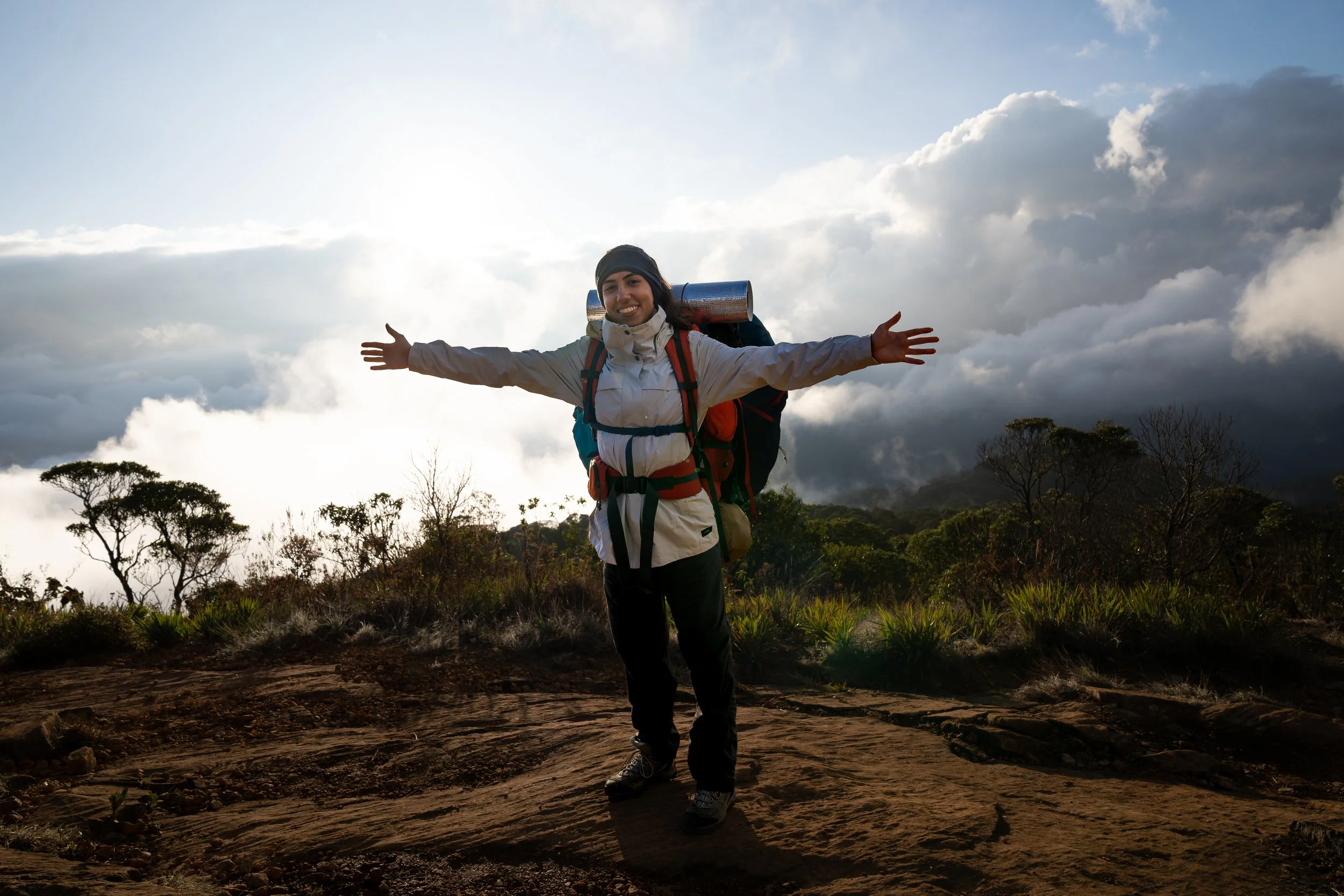 A hiker, Julia Pinna with open arms and a backpack standing on a mountain with a cloudy sky and vegetation in the background.