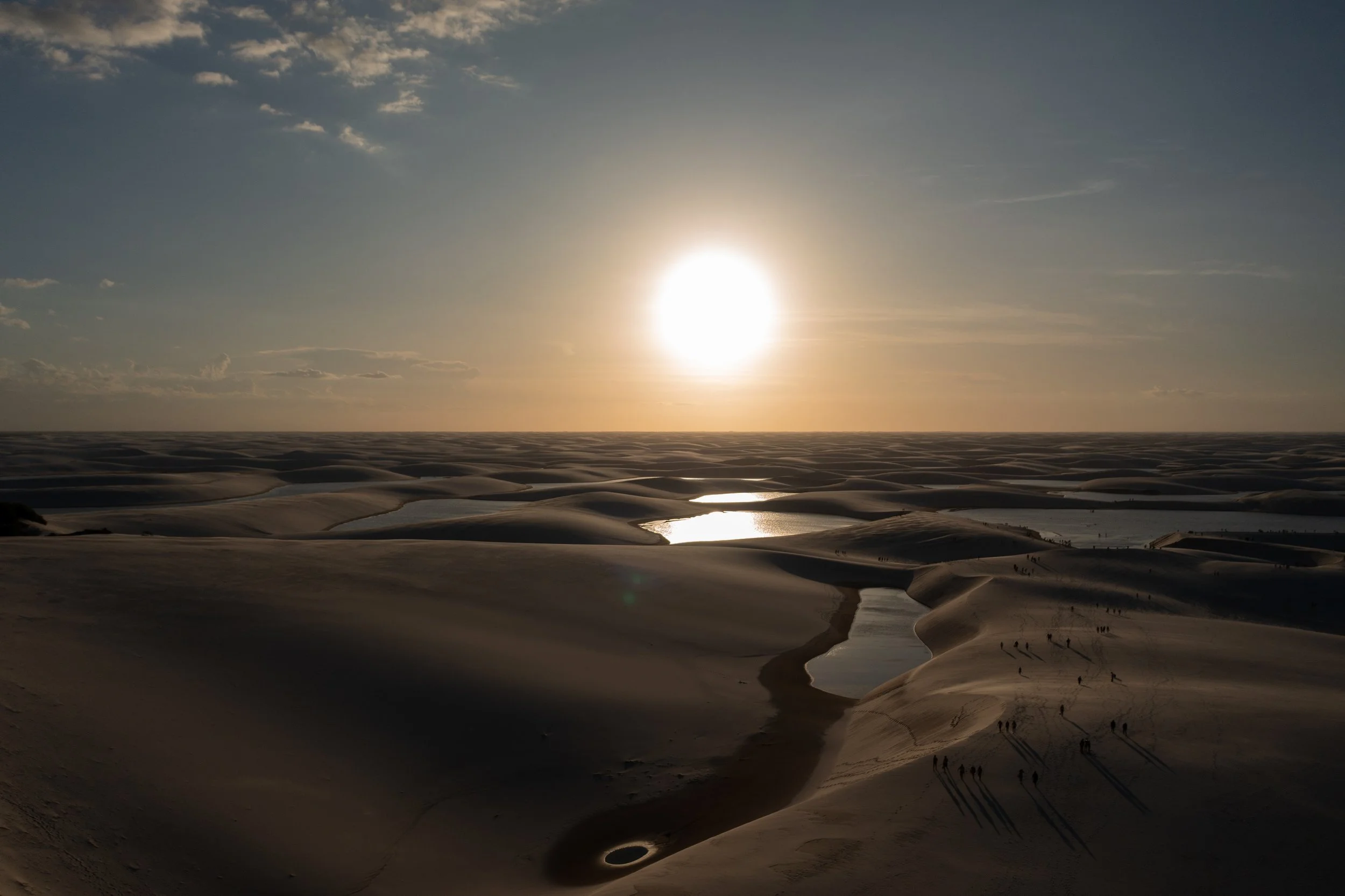 Sand dunes with small water bodies and a setting sun in the desert, with several people walking and casting long shadows.