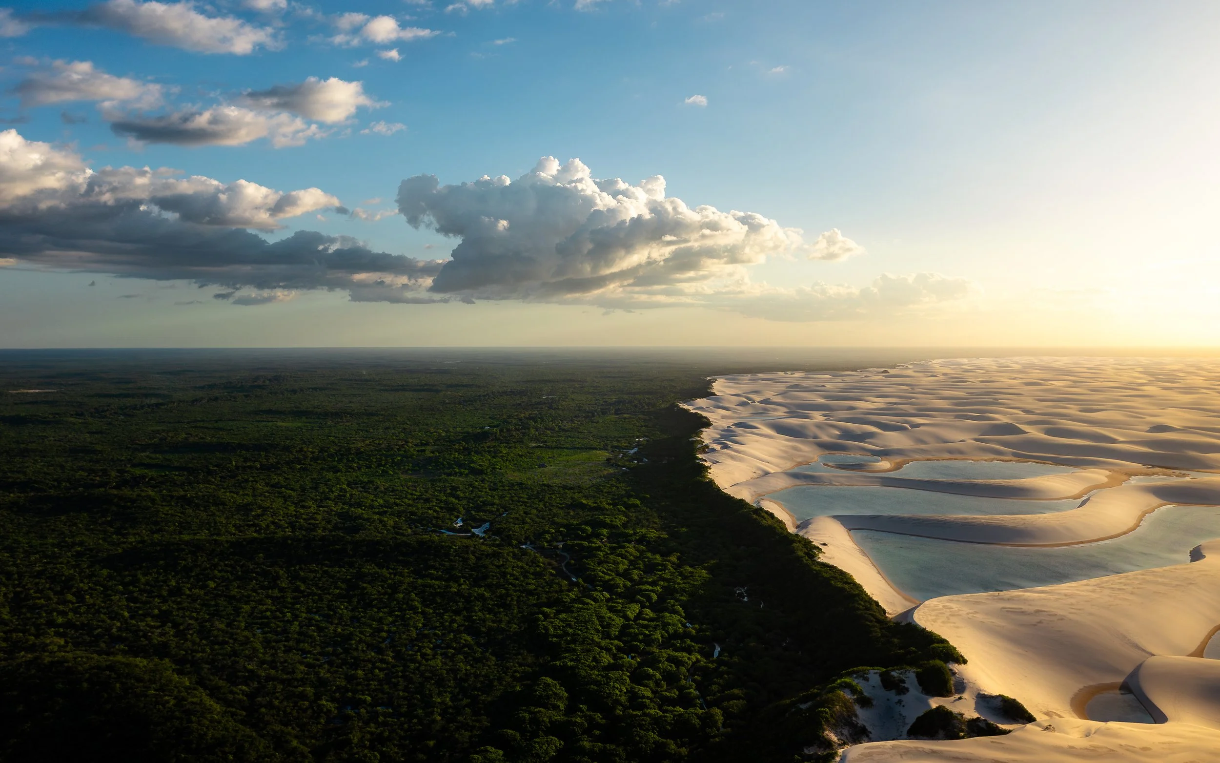 Aerial view of a landscape showing lush green forest on the left and sand dunes with water on the right under a partly cloudy sky.