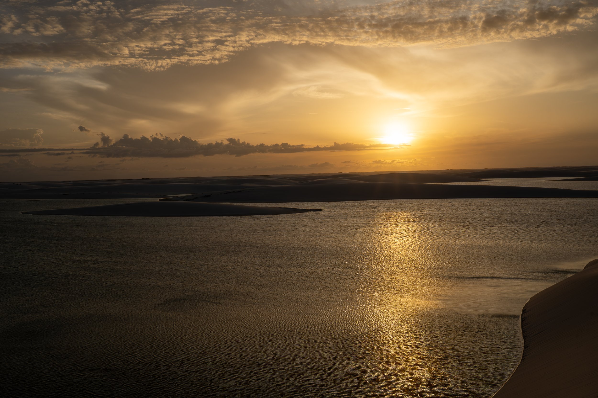 Sunset over sand dunes and a body of water, with clouds in the sky