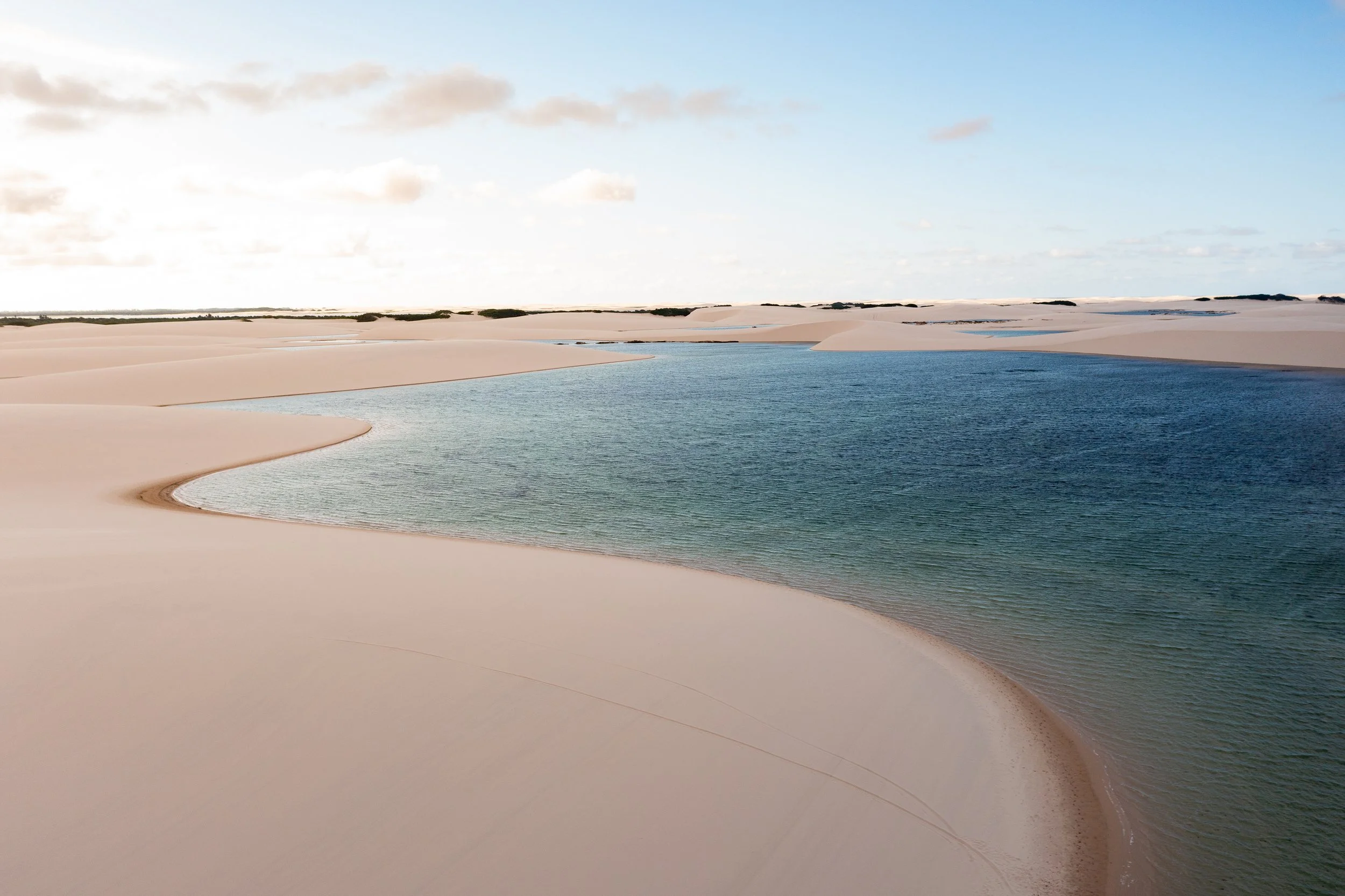 A natural landscape featuring a body of water surrounded by sand dunes and a blue sky with clouds.