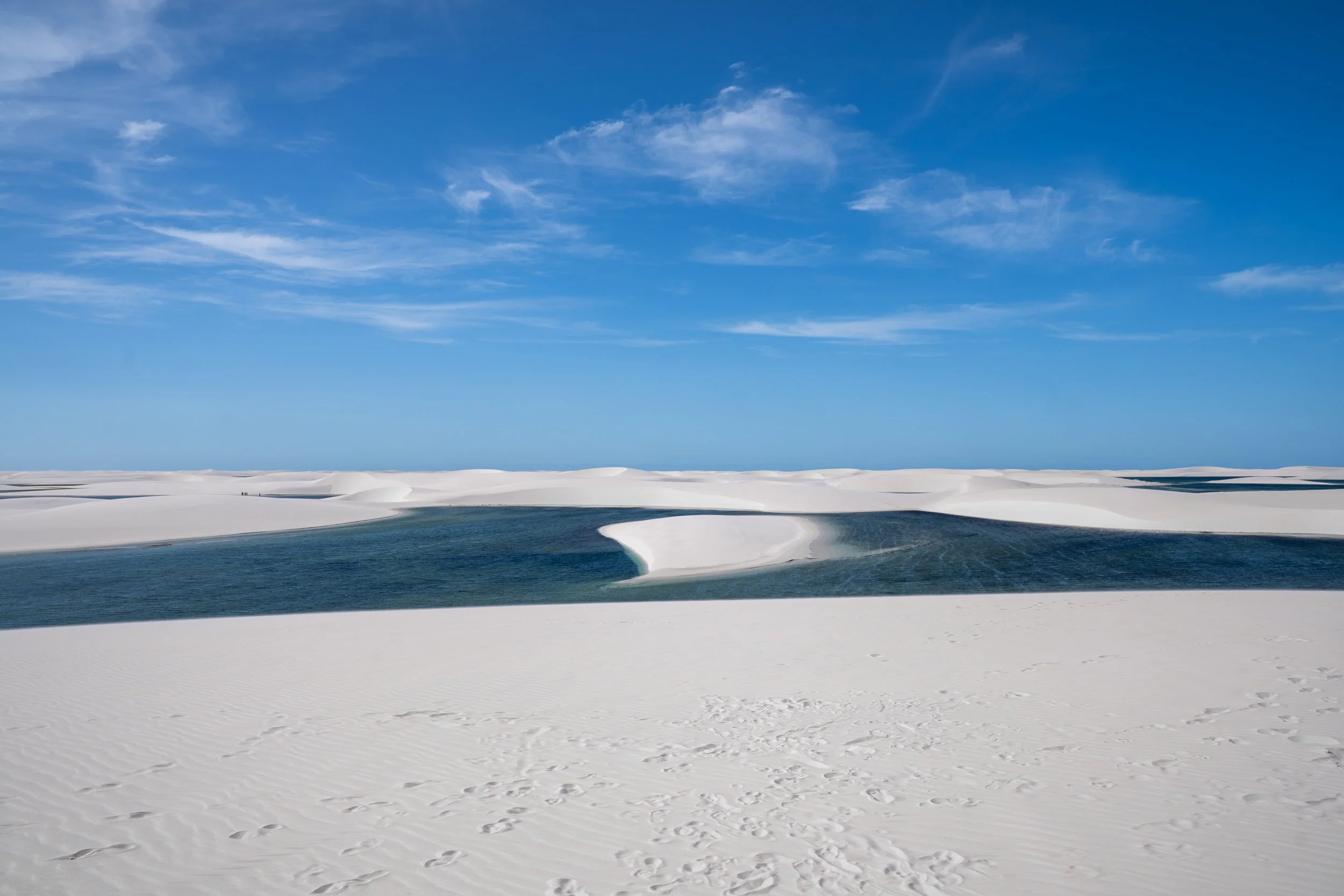 White sand dunes with a blue water lagoon under a clear blue sky with scattered clouds.