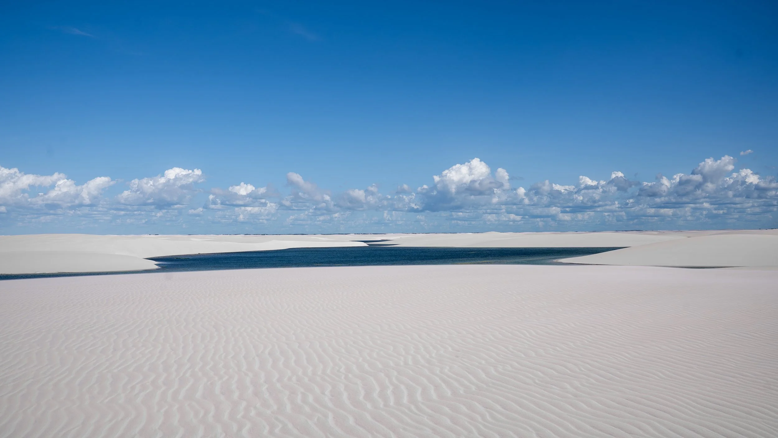 White sand dunes with a small body of water and a blue sky with scattered clouds in the background.