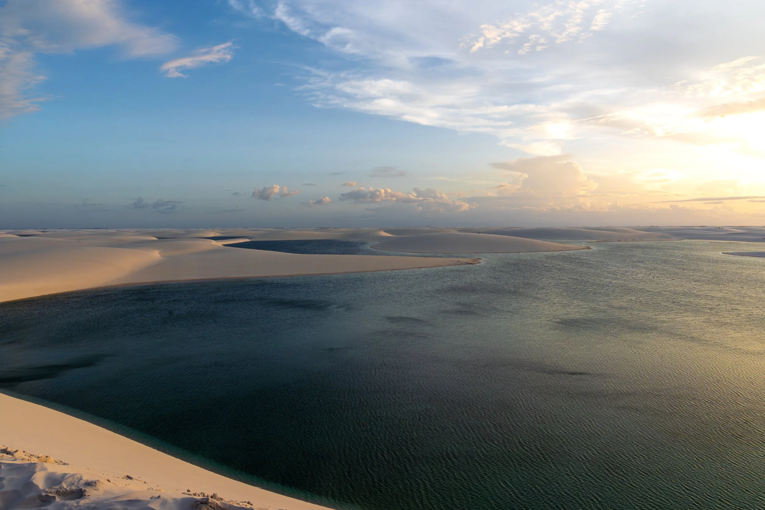 Dunes with a body of water, possibly a lagoon, under a partly cloudy sky during sunset.