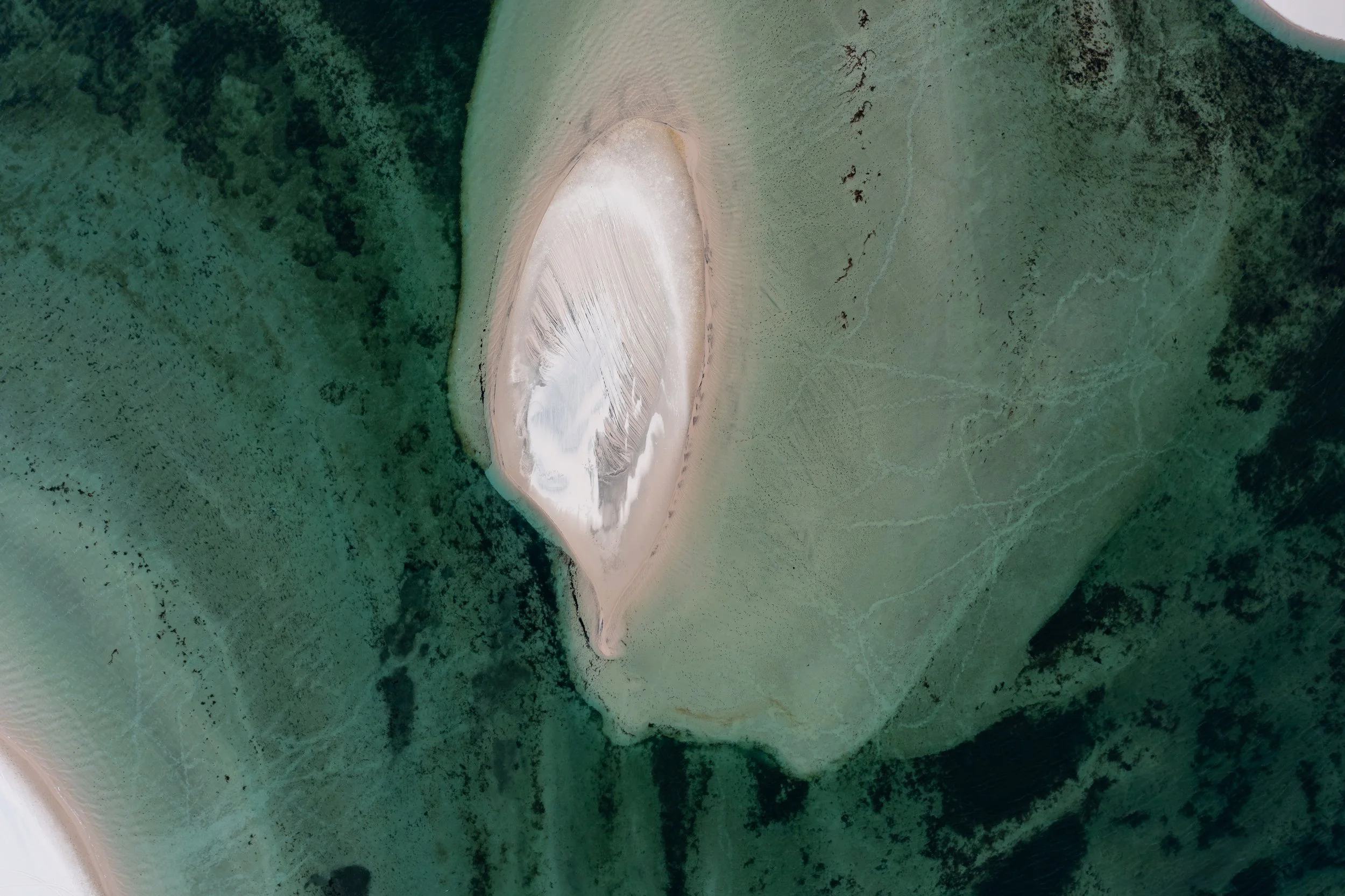 Aerial view of a land formation with a prominent, oval, white sand or salt deposit surrounded by green vegetation and water bodies.