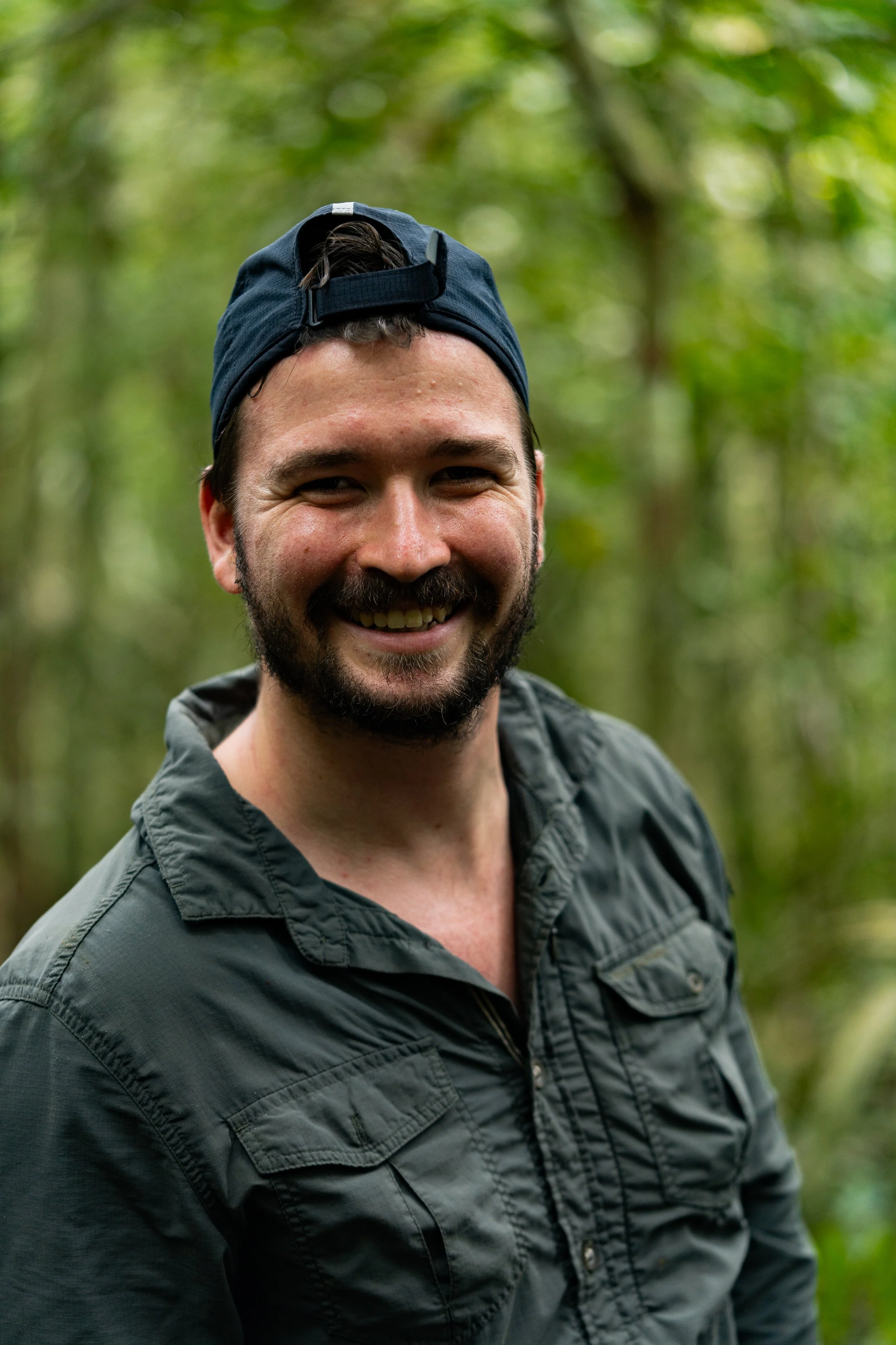 Basil Minder with a beard and cap stands in a forest, smiling.
