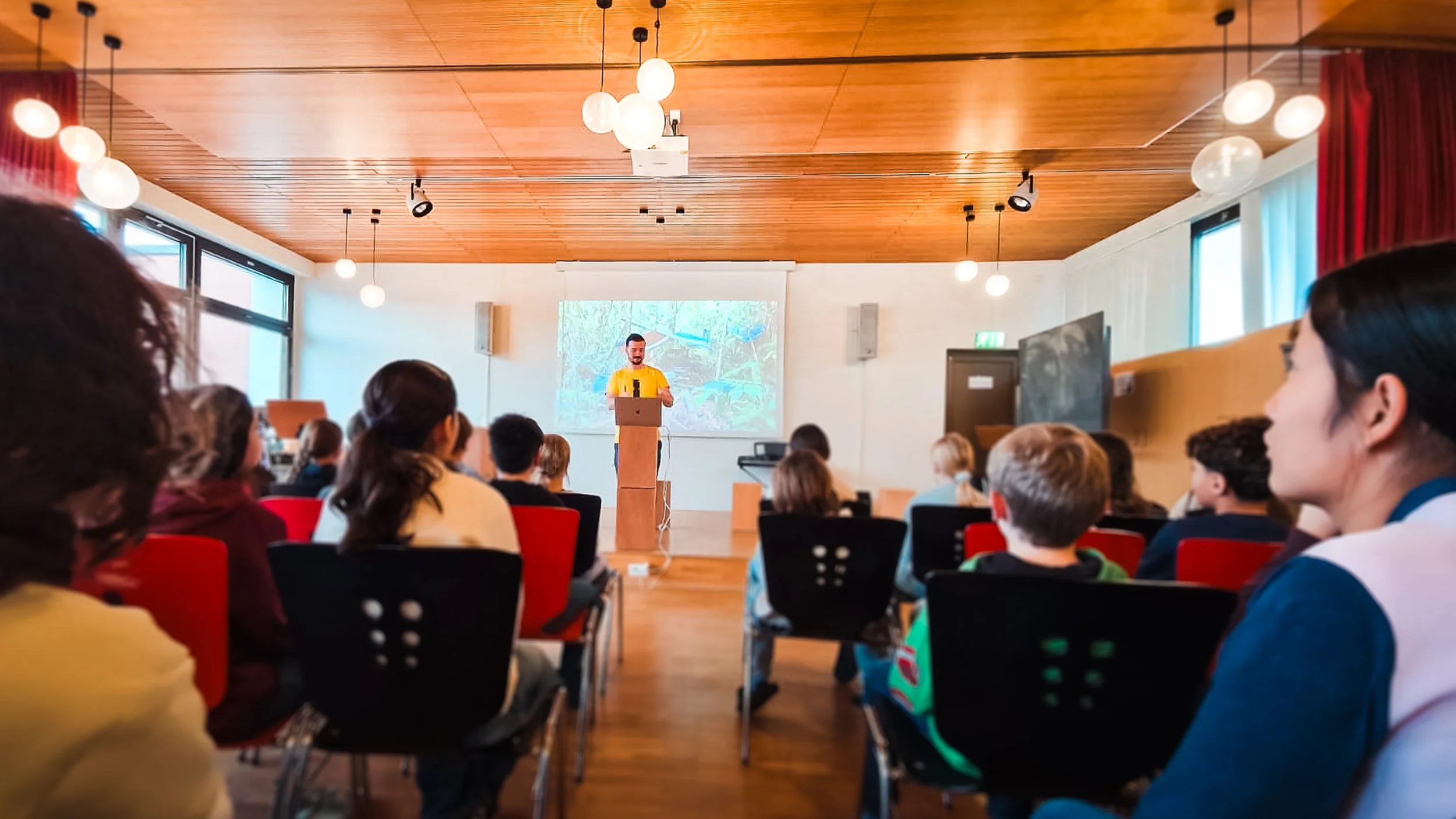 A classroom with children sitting on chairs, facing a man standing at a podium giving a presentation. A projection screen behind him shows an outdoor scene with trees and foliage. The room has wooden ceilings and large windows letting in natural light.