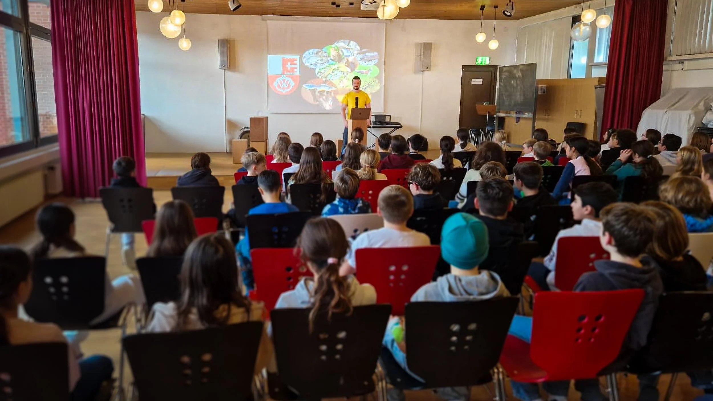 A classroom auditorium filled with children seated and watching a man presenting at a podium on a stage, with a projected colorful graphic behind him.