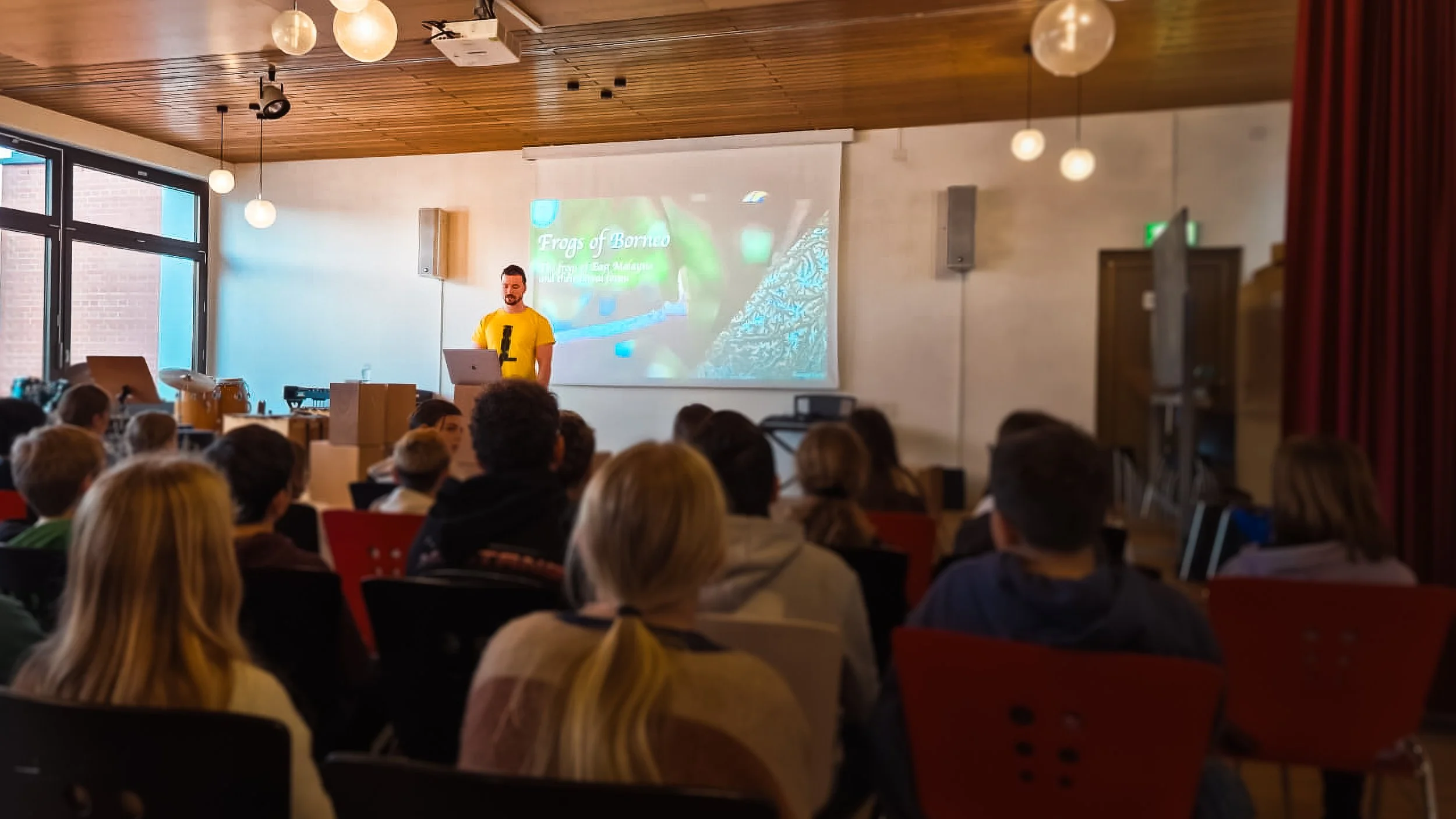 A man wearing a yellow shirt presenting in front of a screen with a slide titled 'Frogs of Borneo' at a lecture or conference with an audience seated in front.