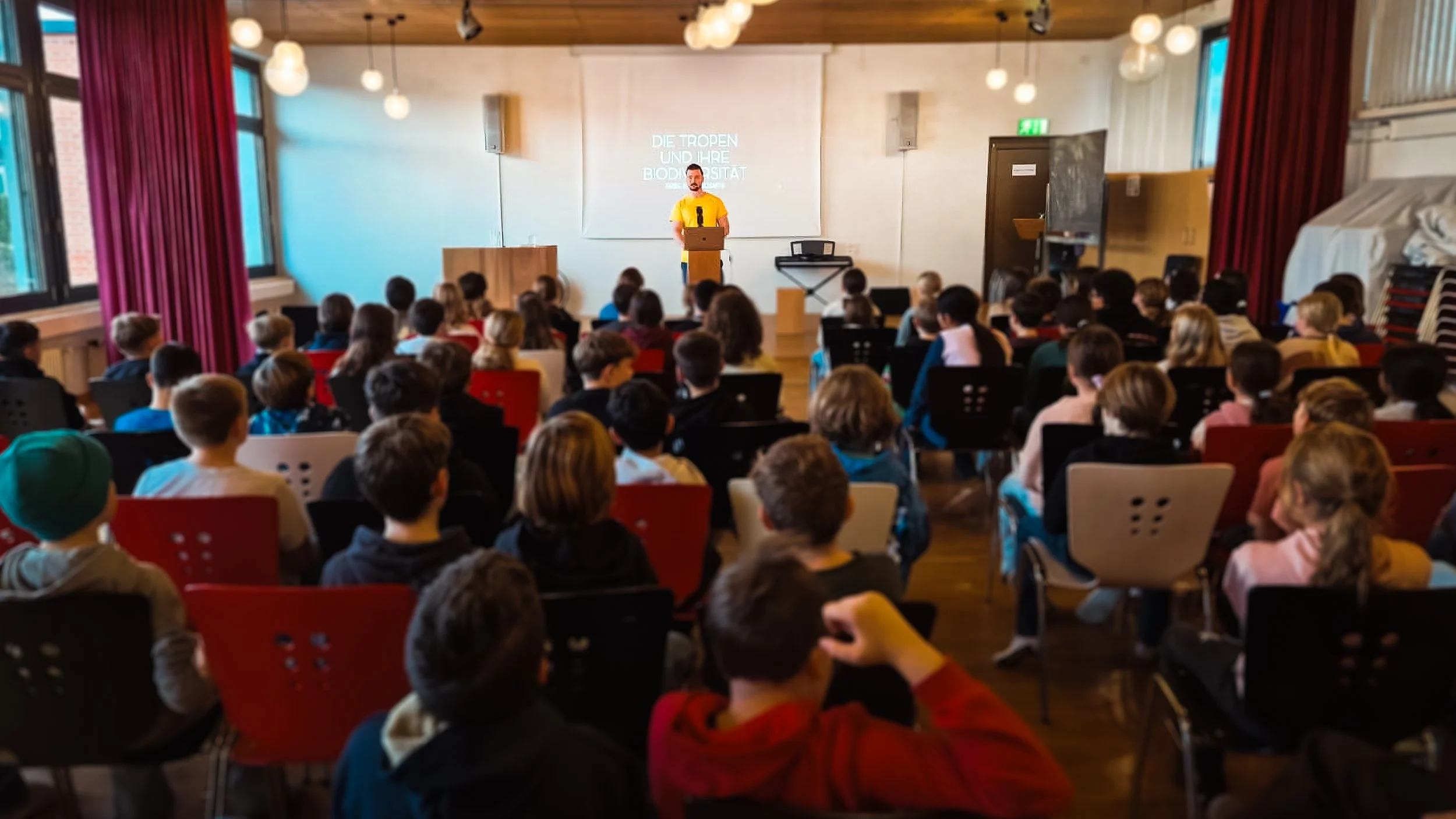 A man giving a presentation to a large group of children in a classroom or auditorium setting, with a projection screen behind him displaying text in German.
