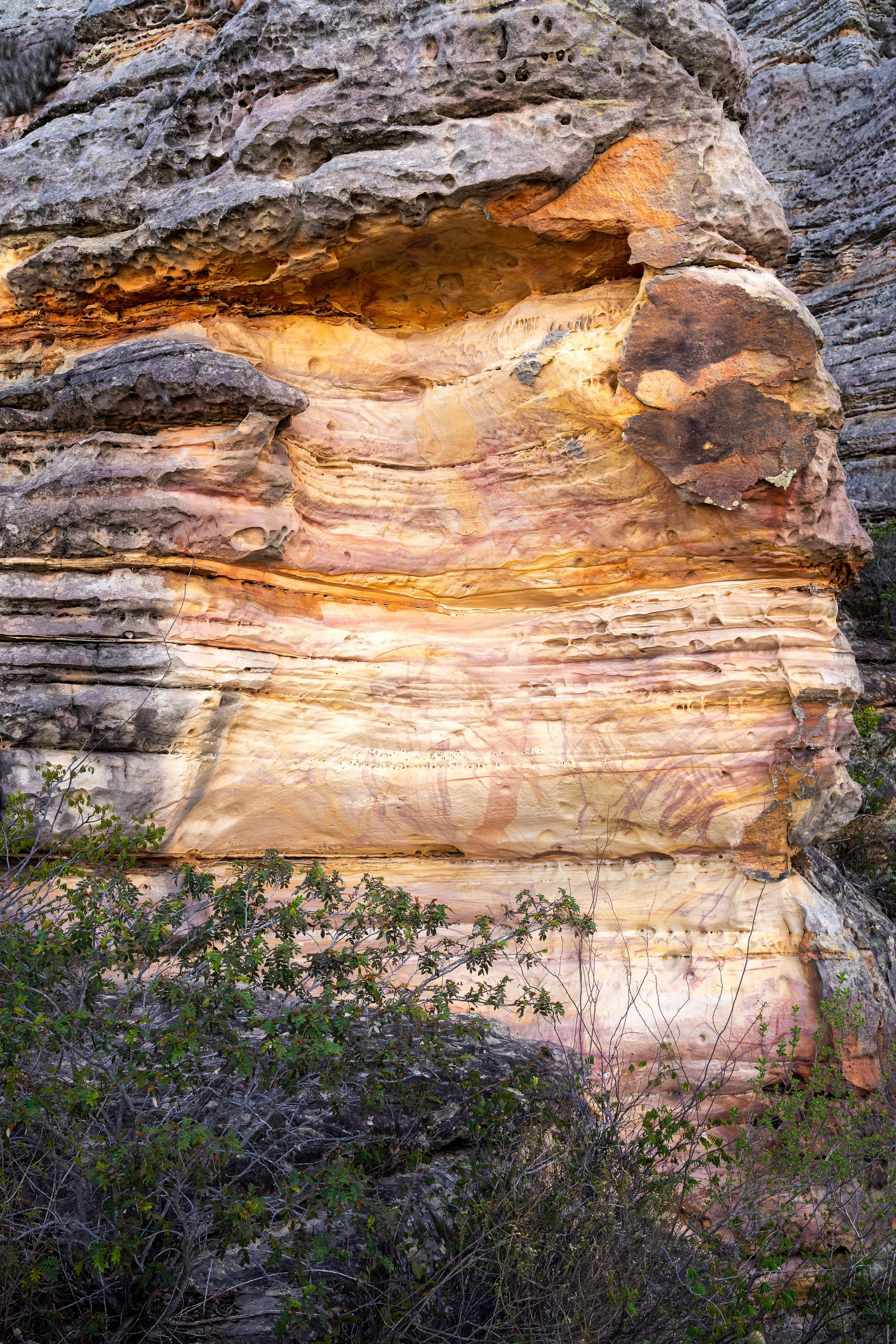Colorful layered rock formation with shades of pinks, yellows, and browns, with some bushes at the bottom.