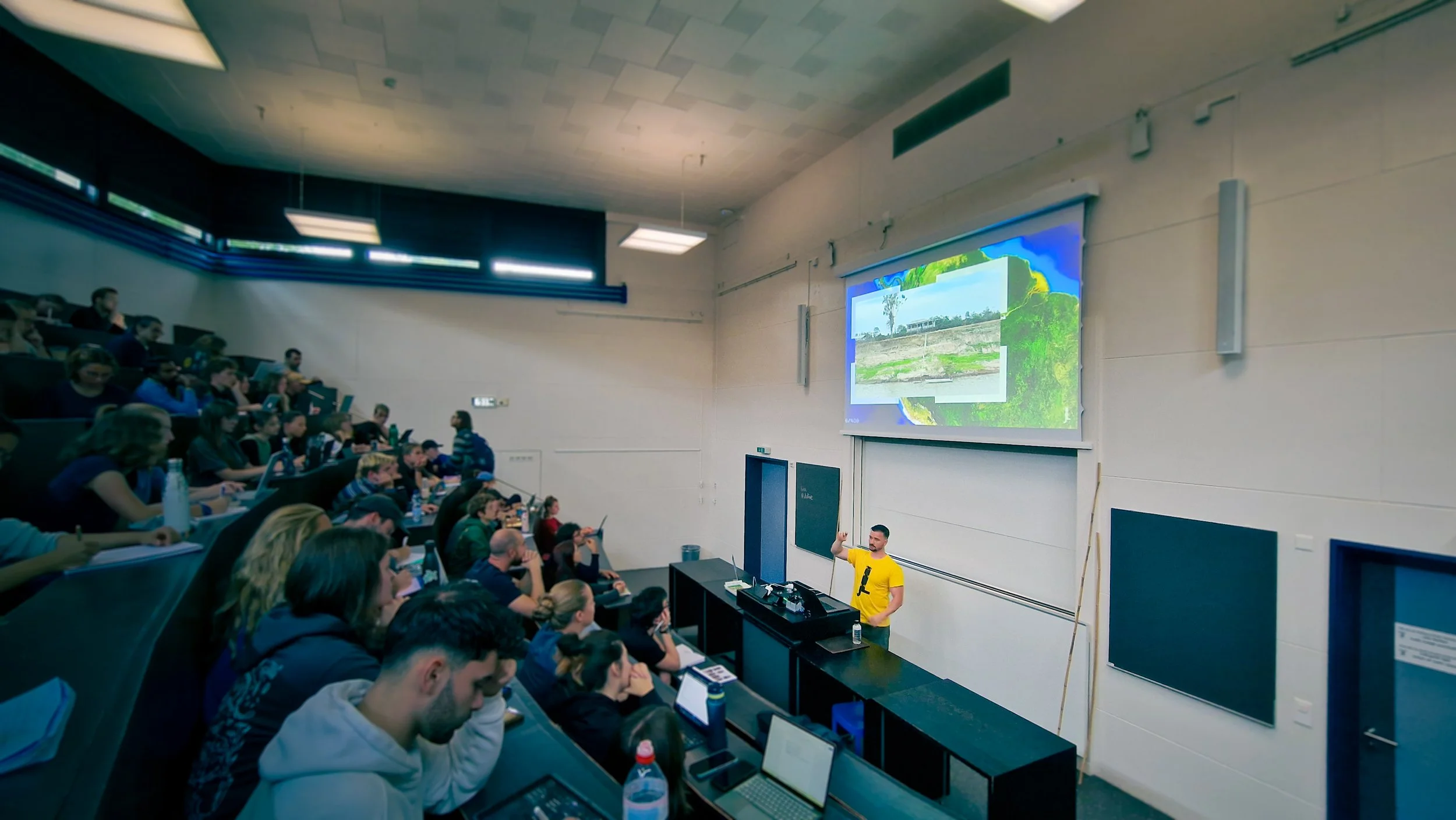 A lecture hall filled with students listening to a presenter giving a talk, with a large screen displaying slides behind him.