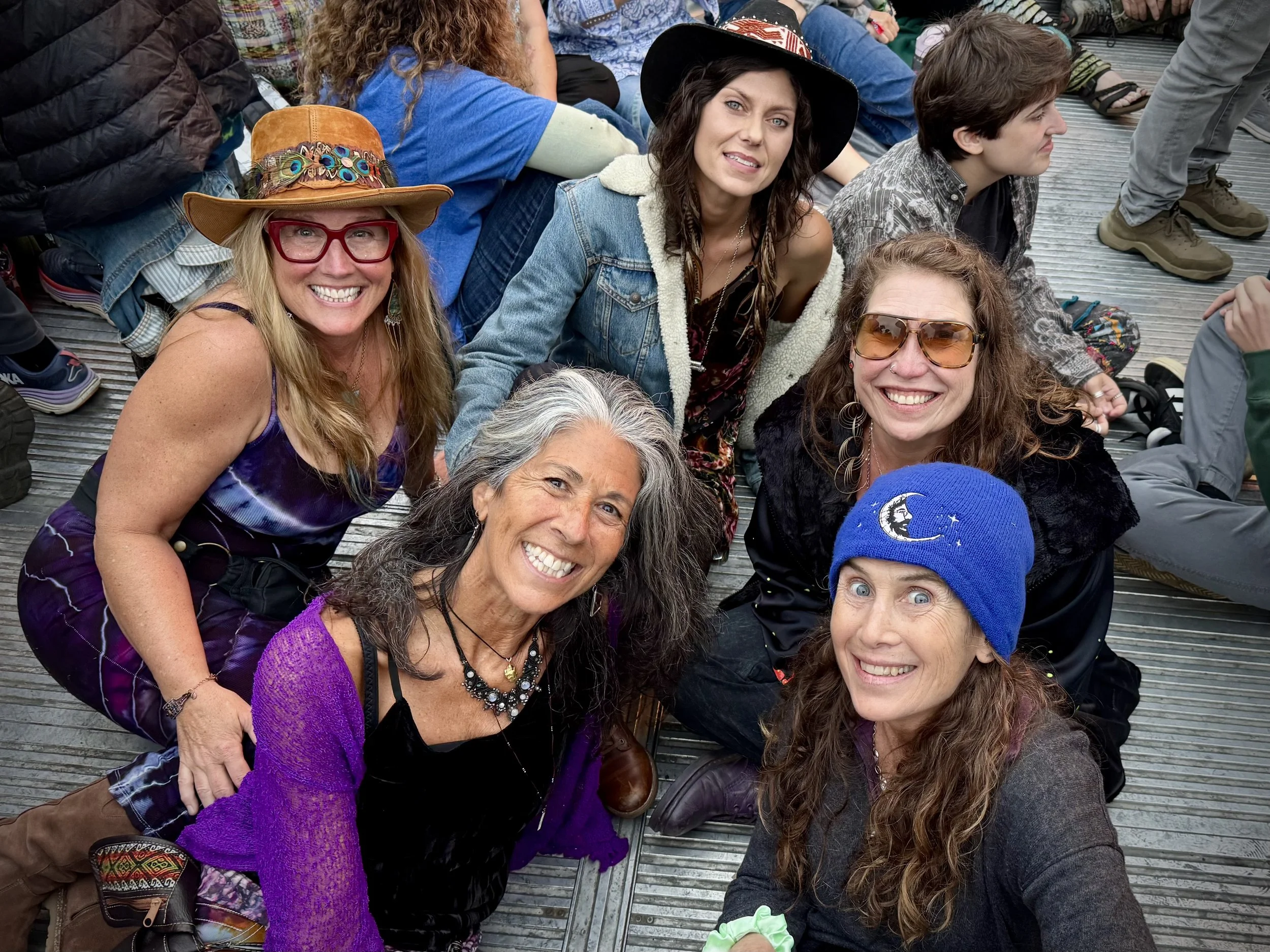 Group of six women sitting and kneeling on a wooden platform, smiling at the camera during an outdoor event. The women have long hair, and some wear glasses, hats, and colorful clothing.