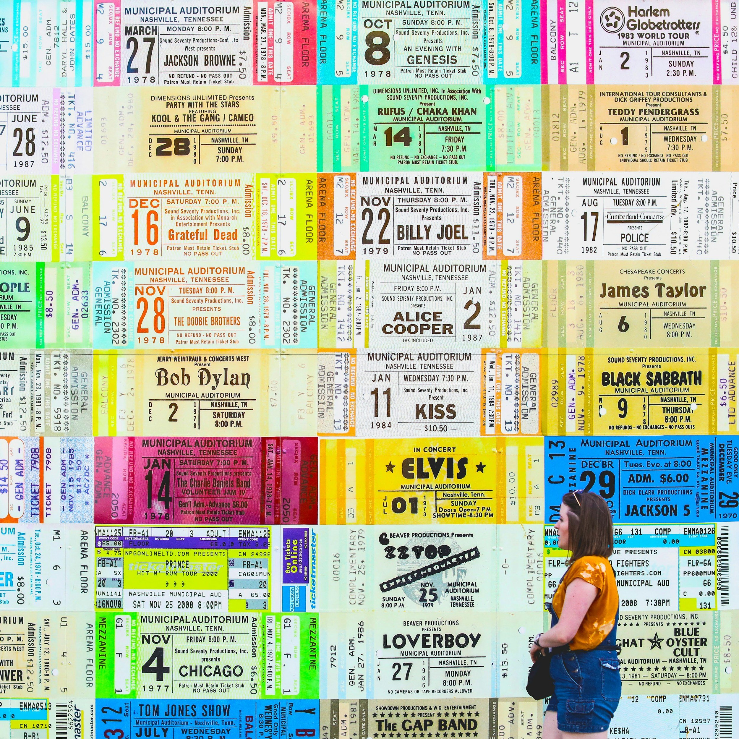 Colorful collage of concert tickets and event stubs arranged in a grid pattern, with a person in a yellow shirt and jeans standing in front at the bottom right corner.