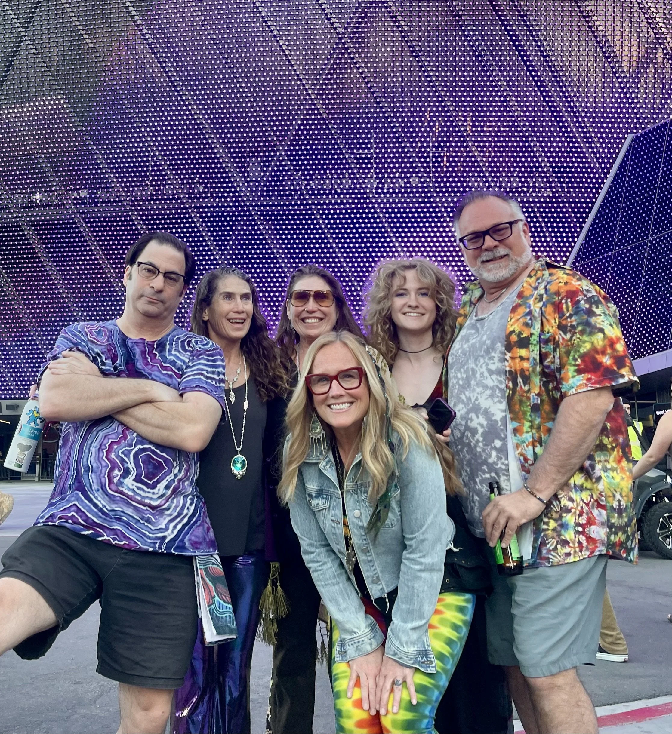 Group of six people taking a photo outdoors in front of a large, colorful LED-lit building at night, dressed in vibrant, casual clothing.