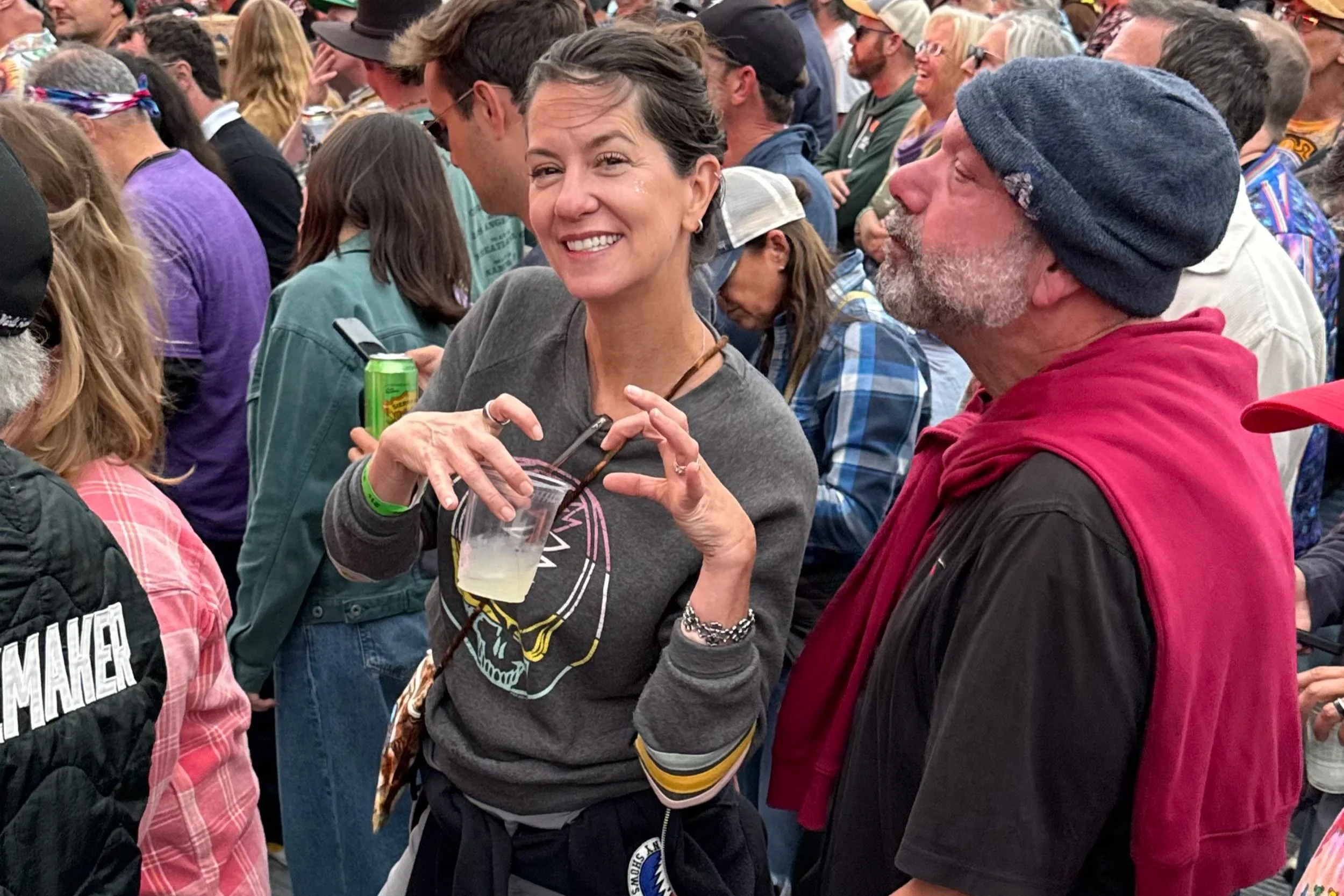 A woman holding a plastic cup with a drink and gesturing with her hand, standing next to a man with a gray beard in a beanie, in a crowd at an outdoor event.