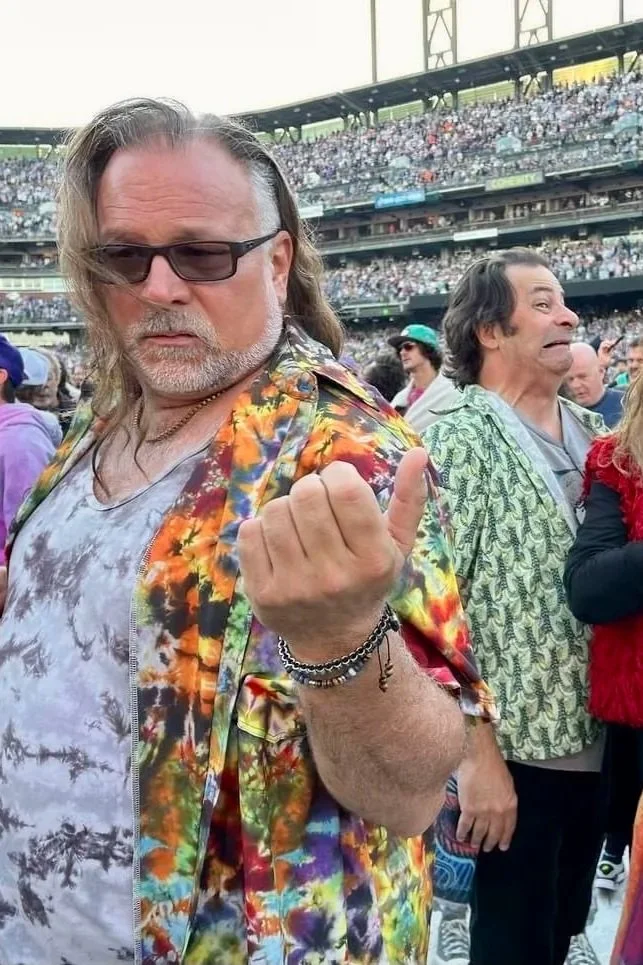 A man with long hair, sunglasses, and a colorful Hawaiian shirt making a fist gesture at a crowded outdoor event with stadium seating in the background.