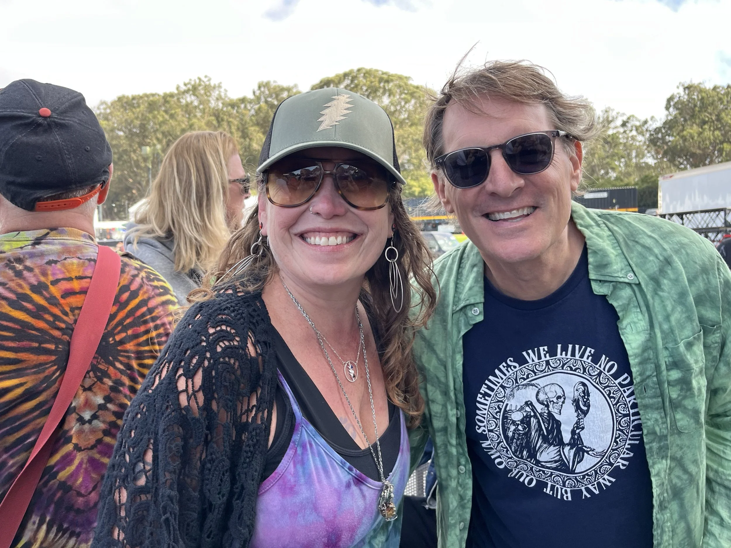 Two smiling adults wearing sunglasses at an outdoor event, with a woman on the left wearing a cap and colorful tie-dye top, and a man on the right wearing a green shirt and black T-shirt.
